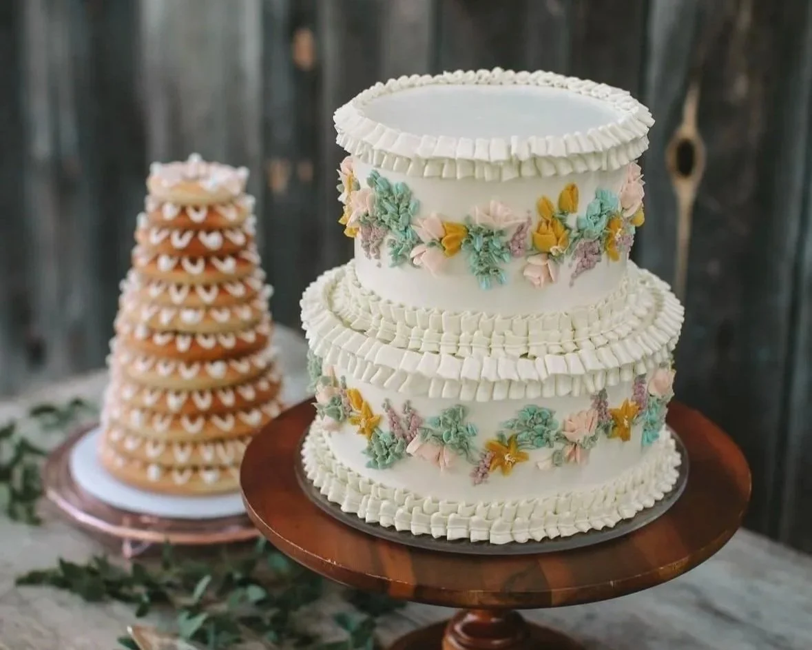 A two-tiered white wedding cake with pastel-colored floral decorations on a wooden cake stand, and a cone-shaped gingerbread house decorated with white icing in the background.