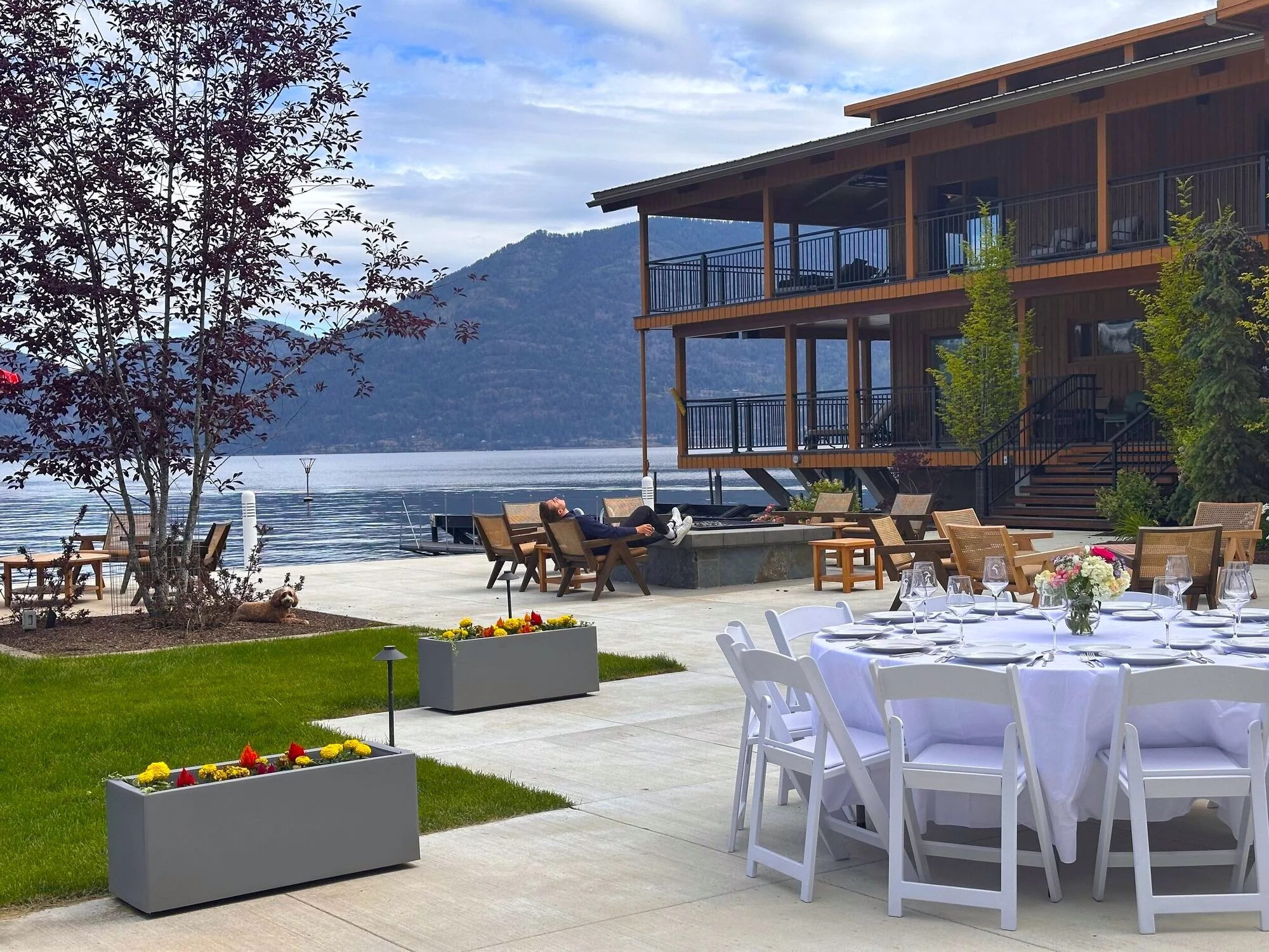 An outdoor lakeside dining area with a large wooden house on stilts, a man relaxing in a chair, yellow flowers in a rectangular planter, white tables with glassware, a tree with reddish leaves, and a background of mountains and a cloudy sky.