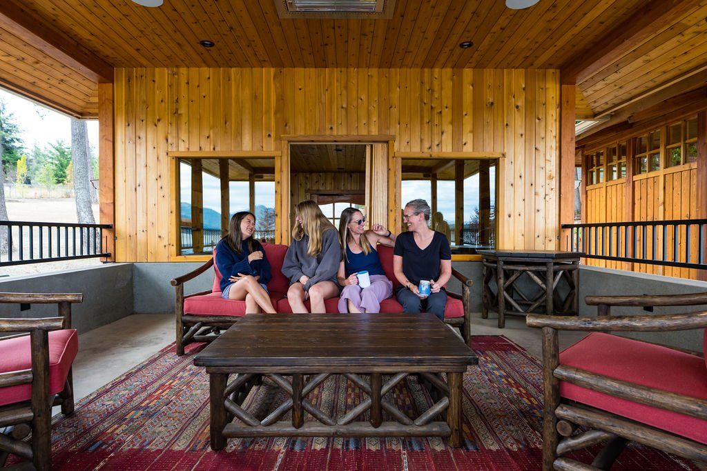 Four women sitting together on a red cushioned outdoor sofa in a wooden porch, engaged in conversation and smiling.