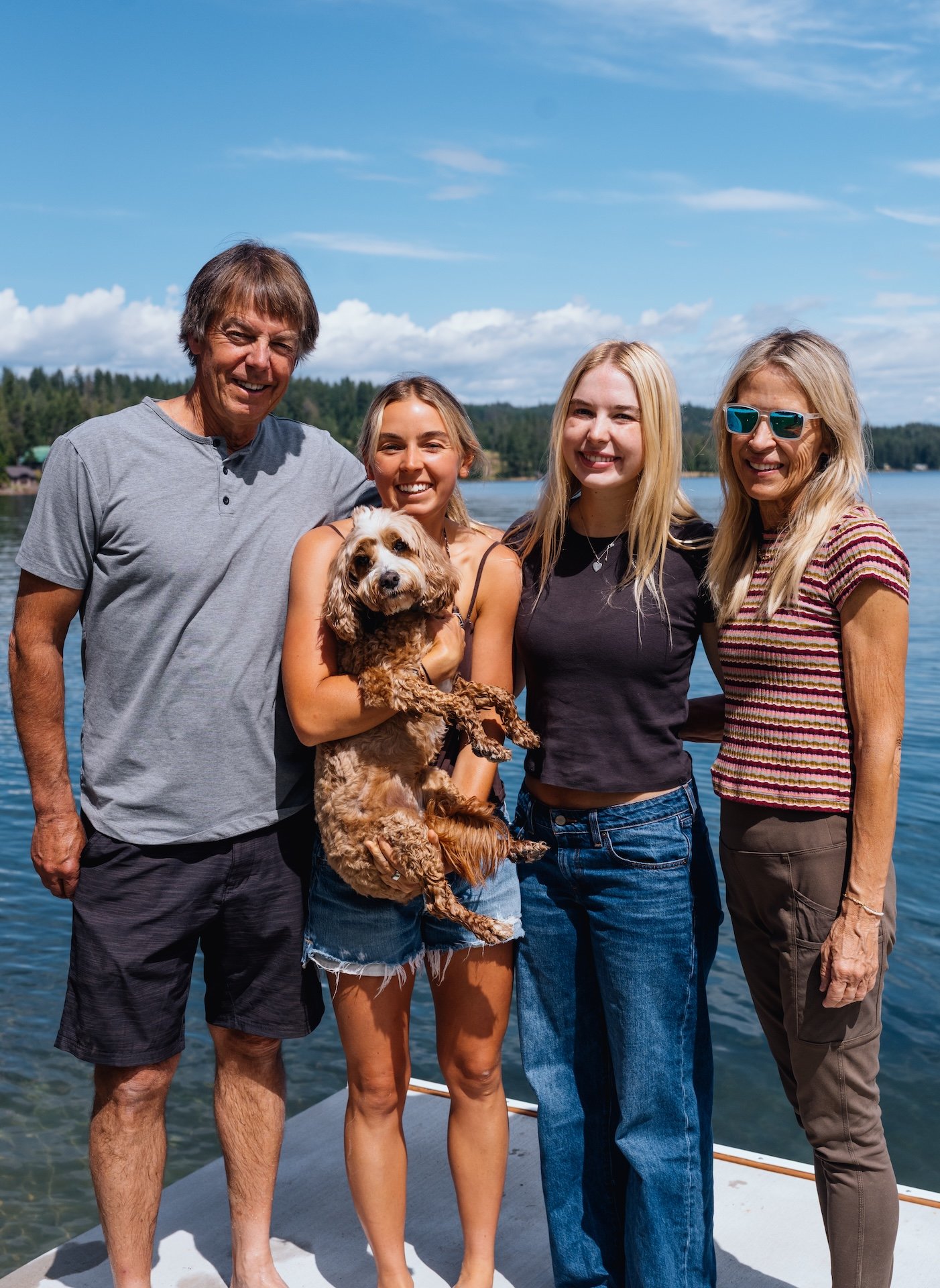A group of four people and a dog standing on a boat near a lake, with trees and blue sky in the background.