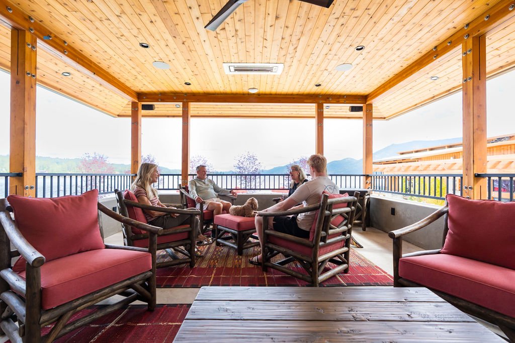 Family sitting and talking on a covered balcony with wooden ceiling, railing, and mountain view in the background.