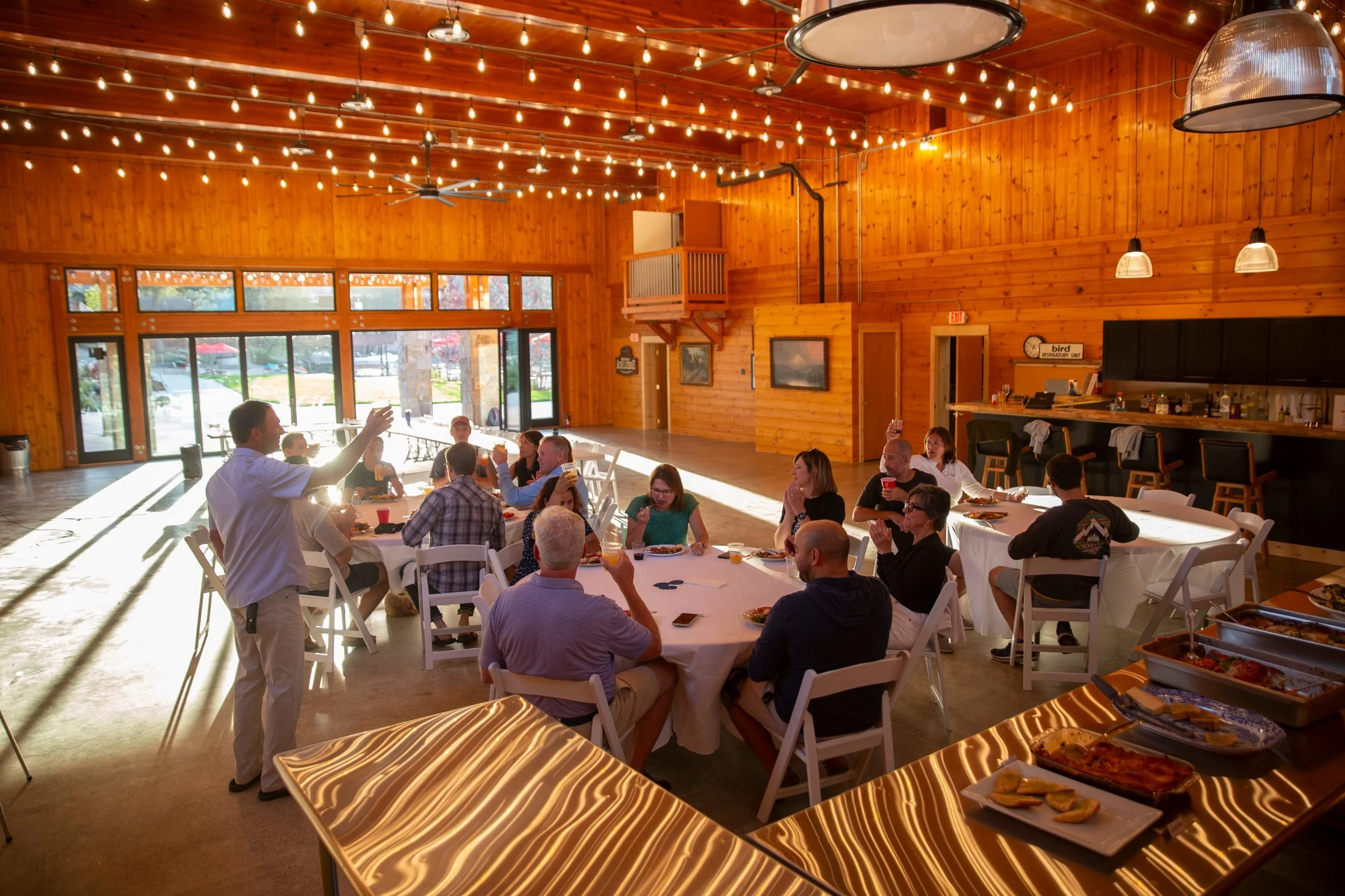 People gathered indoors for a meal at a restaurant with wooden walls and ceiling, string lights, large windows, and buffet food on tables.
