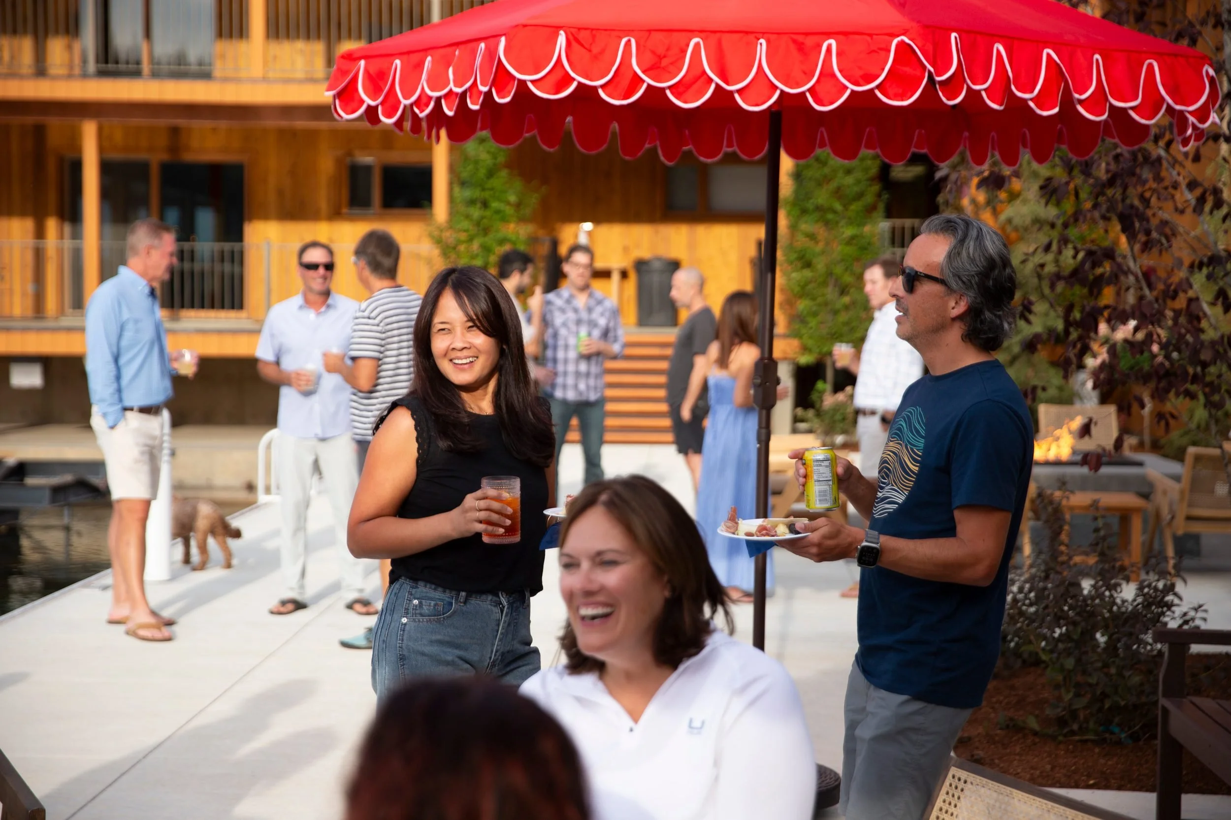 People enjoying a social gathering outside under a red patio umbrella, with wooden buildings and autumn foliage in the background.