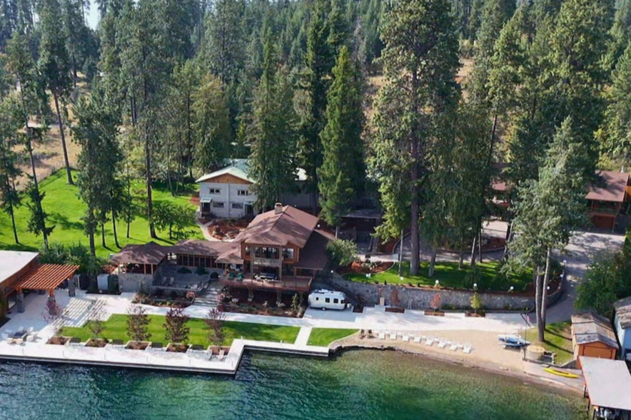 Aerial view of lakeside property with house, dock, boat, trees, and green lawn.
