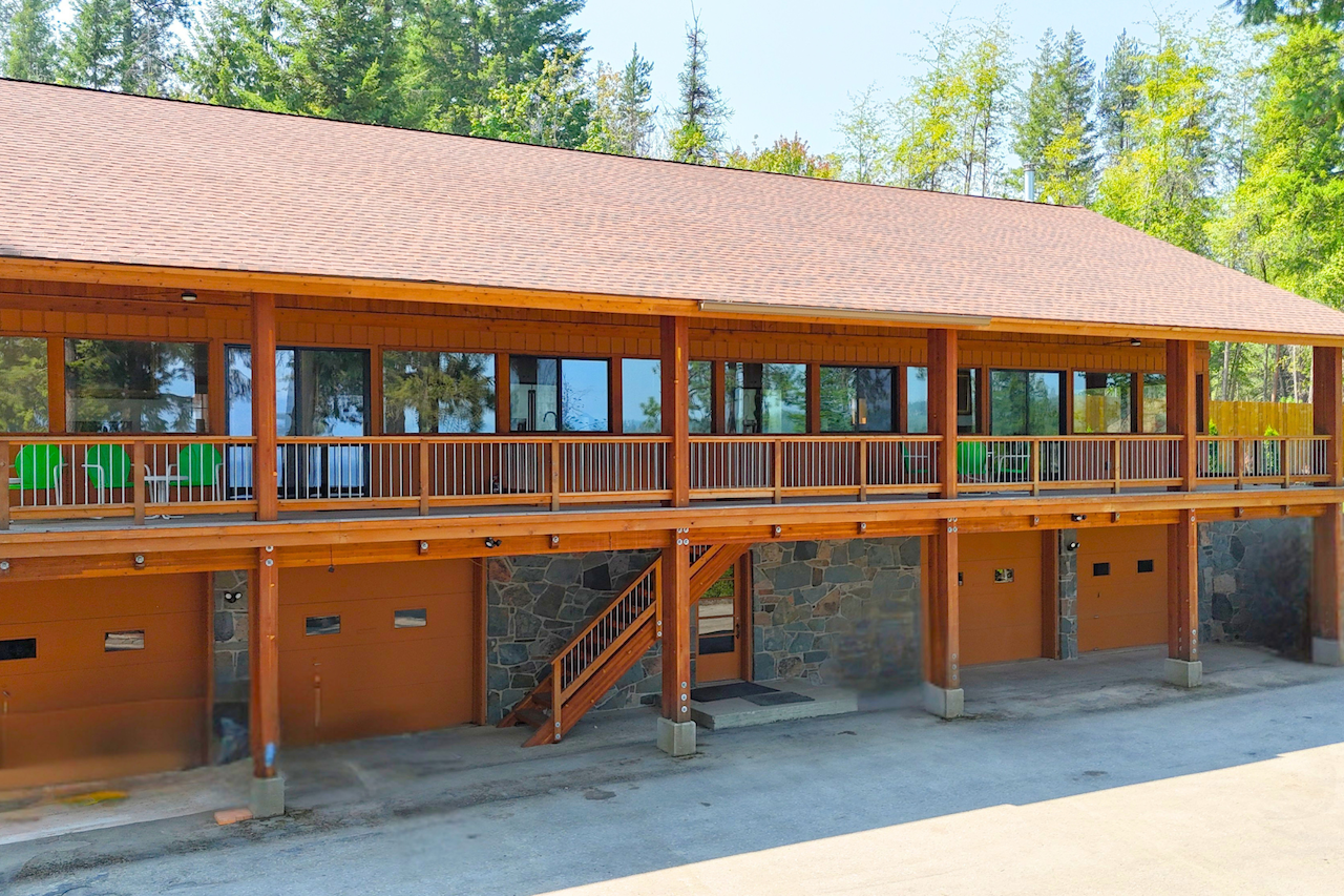 A large wooden house with a spacious balcony on the upper level, surrounded by trees and greenery.
