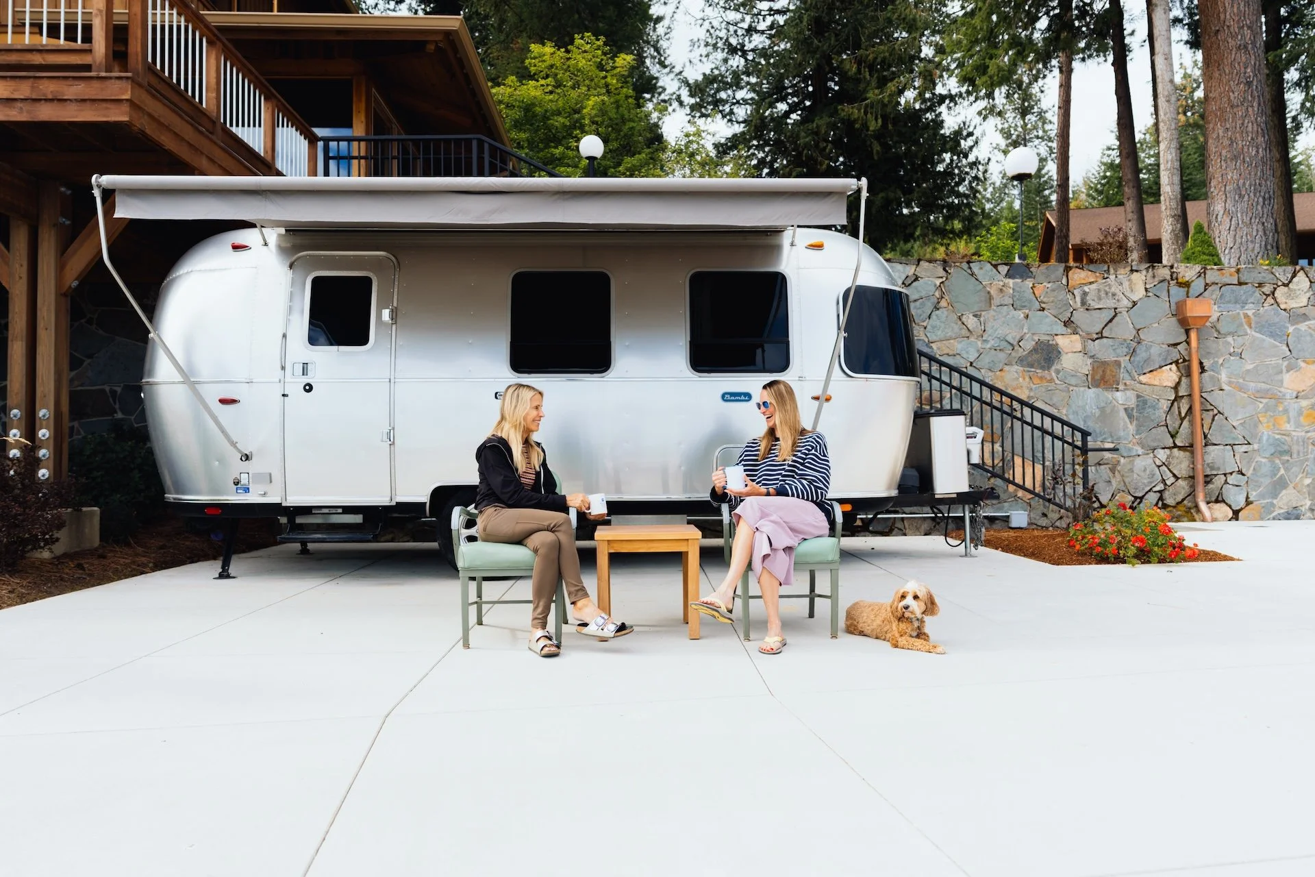 Two women sitting on chairs and chatting in front of a silver camper trailer, with a dog lying on the ground nearby. The scene is outdoors with a stone wall, trees, and a house in the background.