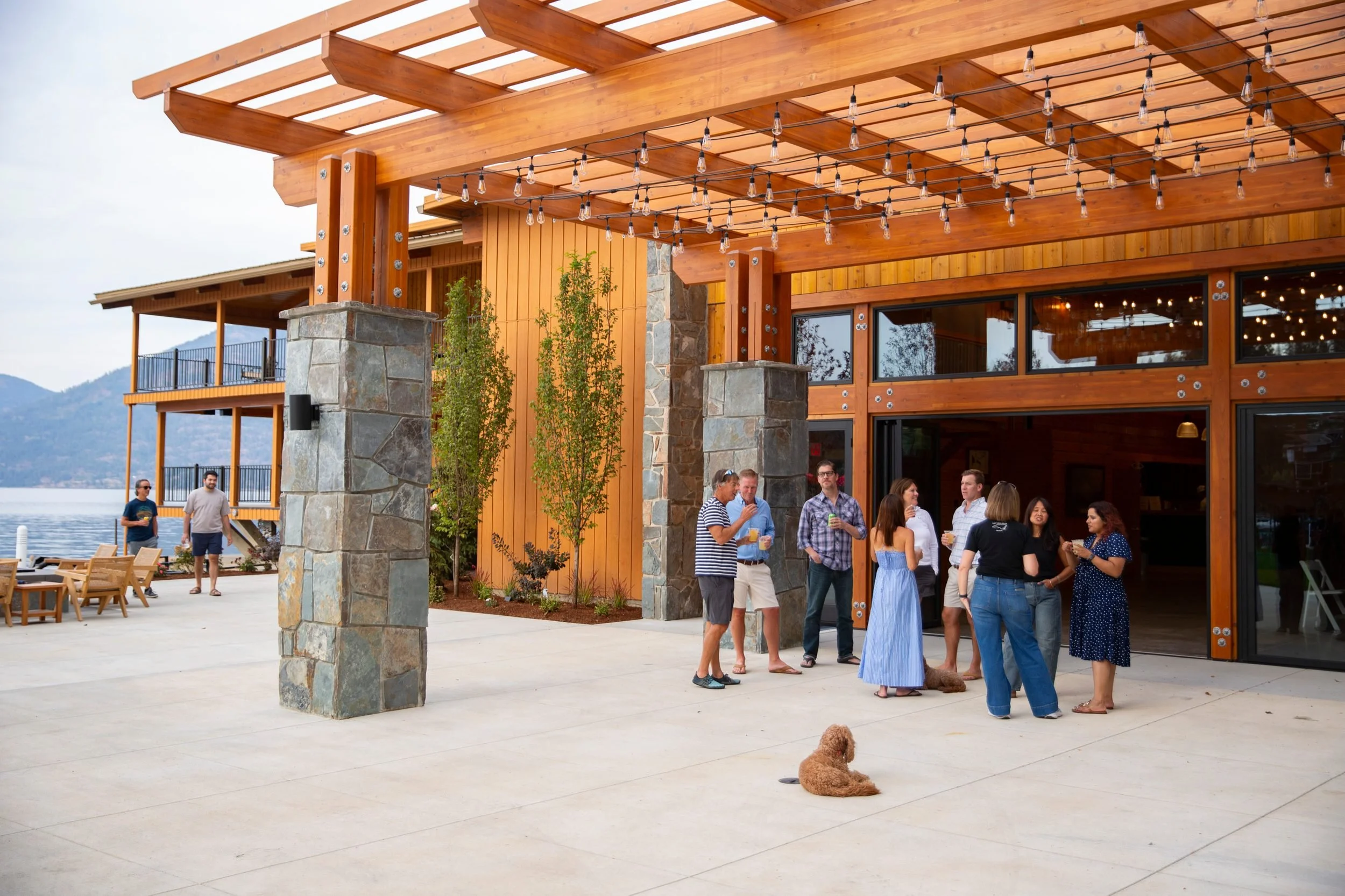 People gathered outdoors at a wooden building with string lights hanging. Some are socializing, and a dog is lying on the ground. Mountains are visible in the background.