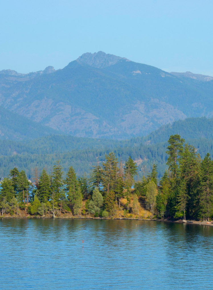 A scenic view of a mountain with a dense forest at its base, next to a calm lake reflecting the trees and sky.