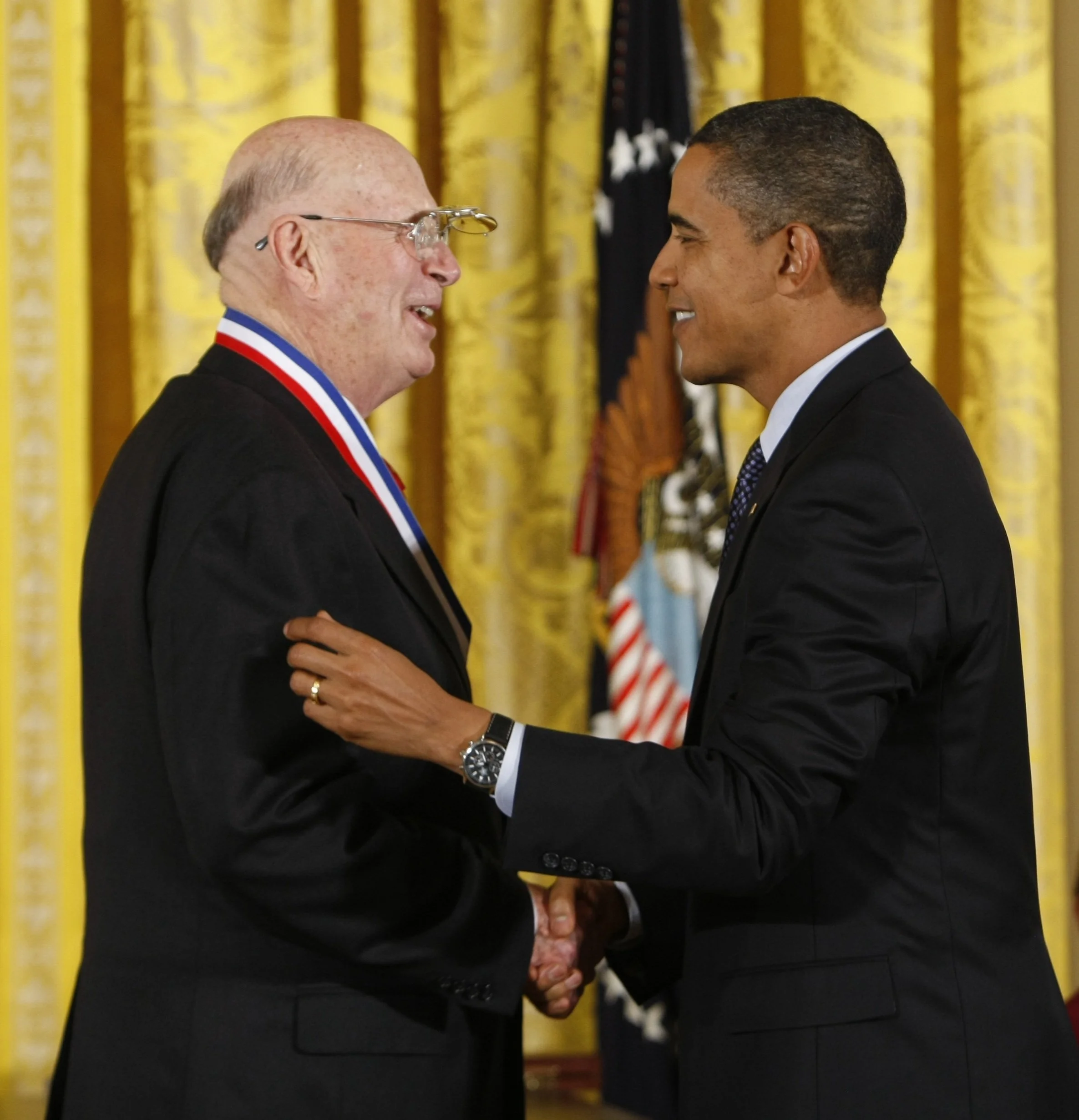 Two men in tuxedos are shaking hands in a formal setting with yellow curtains and flags in the background.