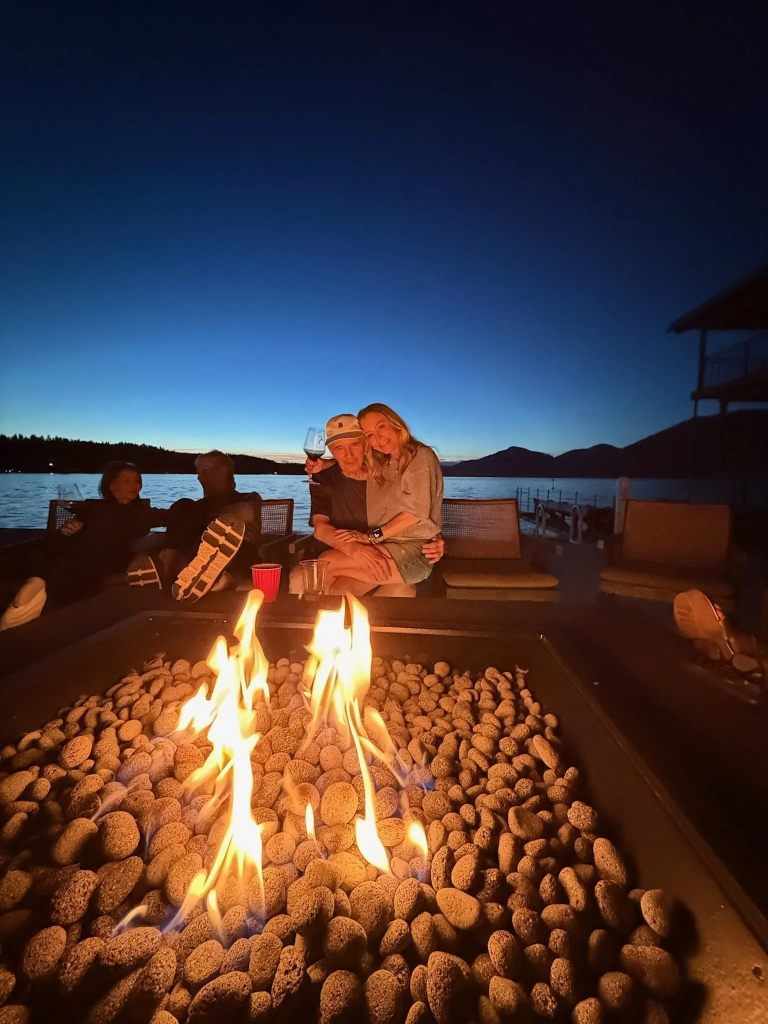 People sitting around a fire pit on a deck overlooking a lake during dusk, with mountains and a house in the background.