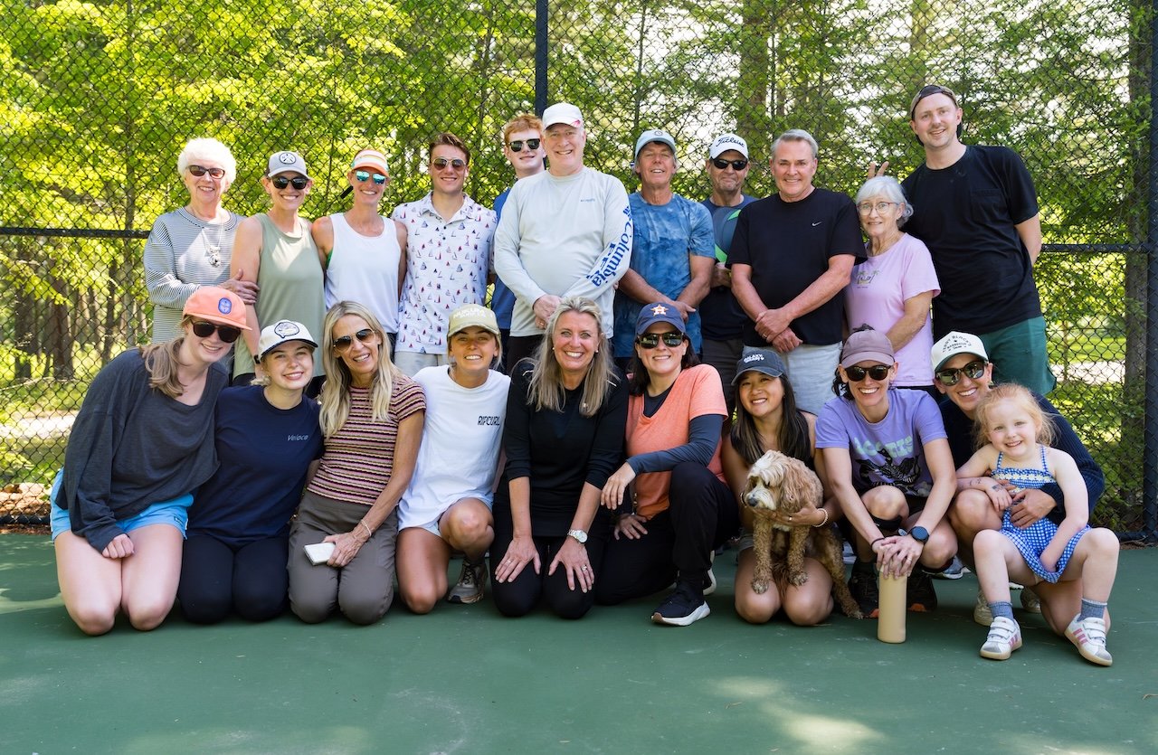 A group of 17 people, including children and adults, posing together outdoors on a tennis court with a chain-link fence and green trees in the background, smiling for the photo.