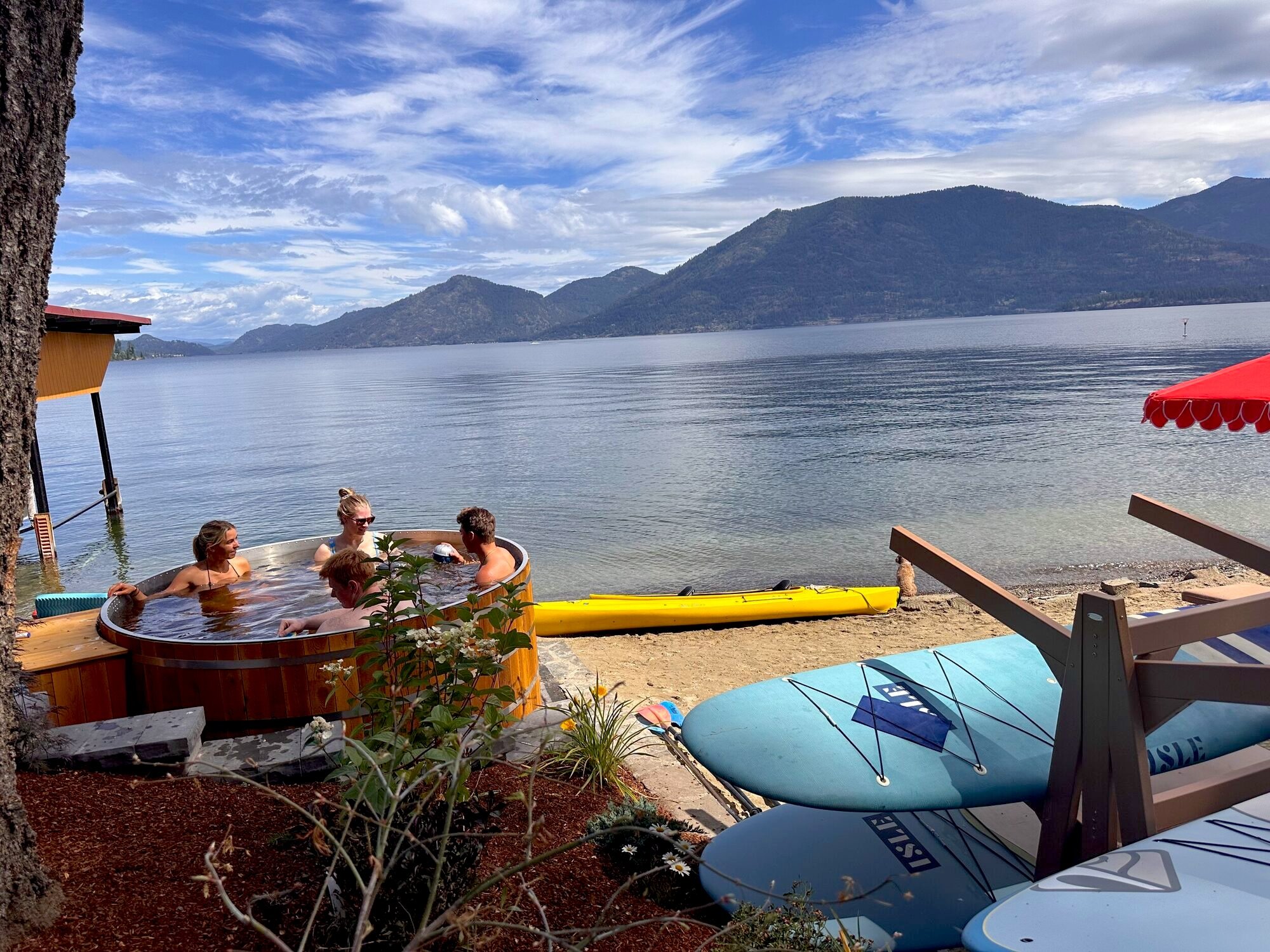 People enjoying a hot tub near a lakeside with mountains in the background on a partly cloudy day.