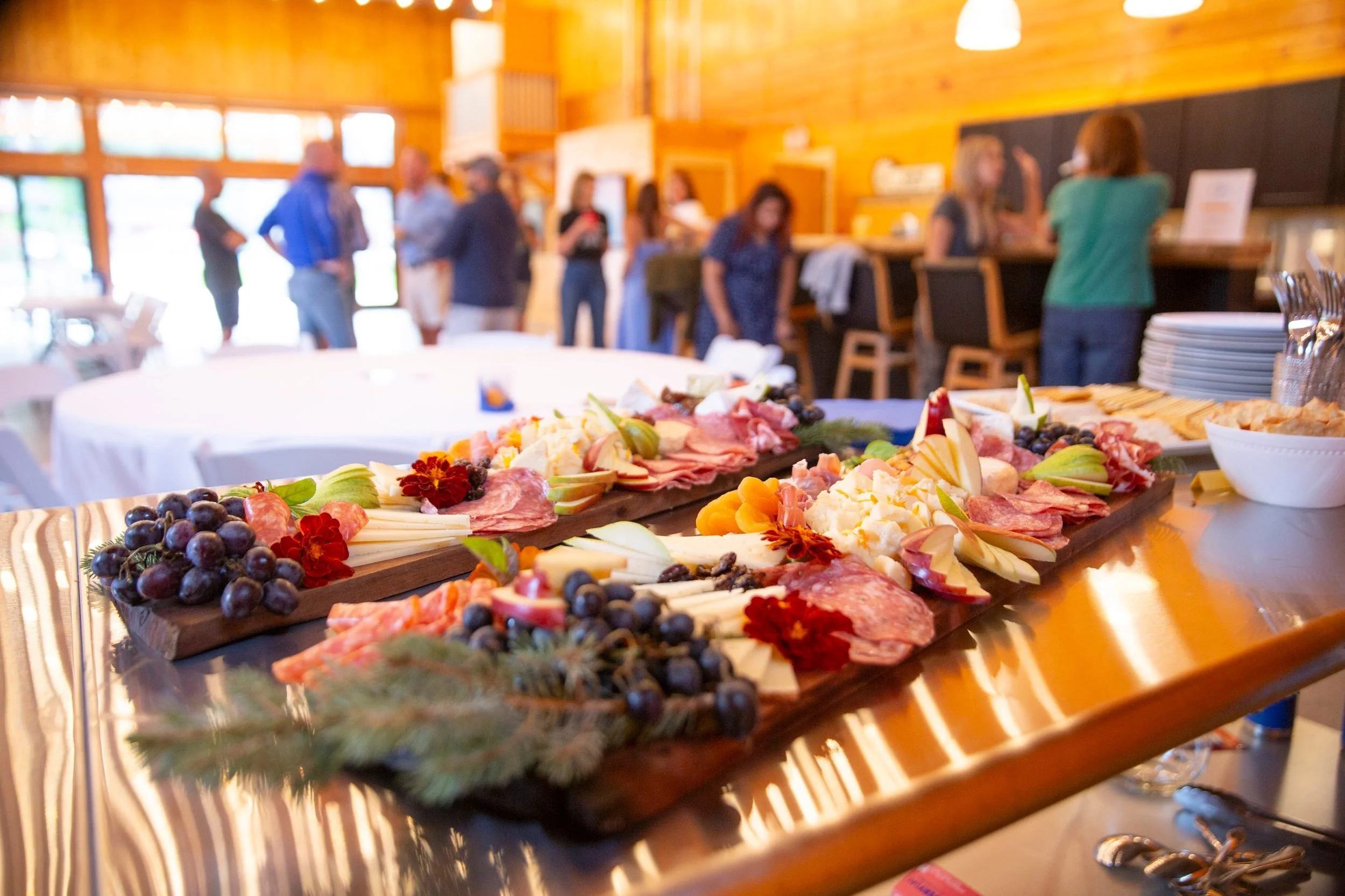 Charcuterie board with grapes, cheeses, meats, and garnishes on a table at a social gathering inside a wooden building.