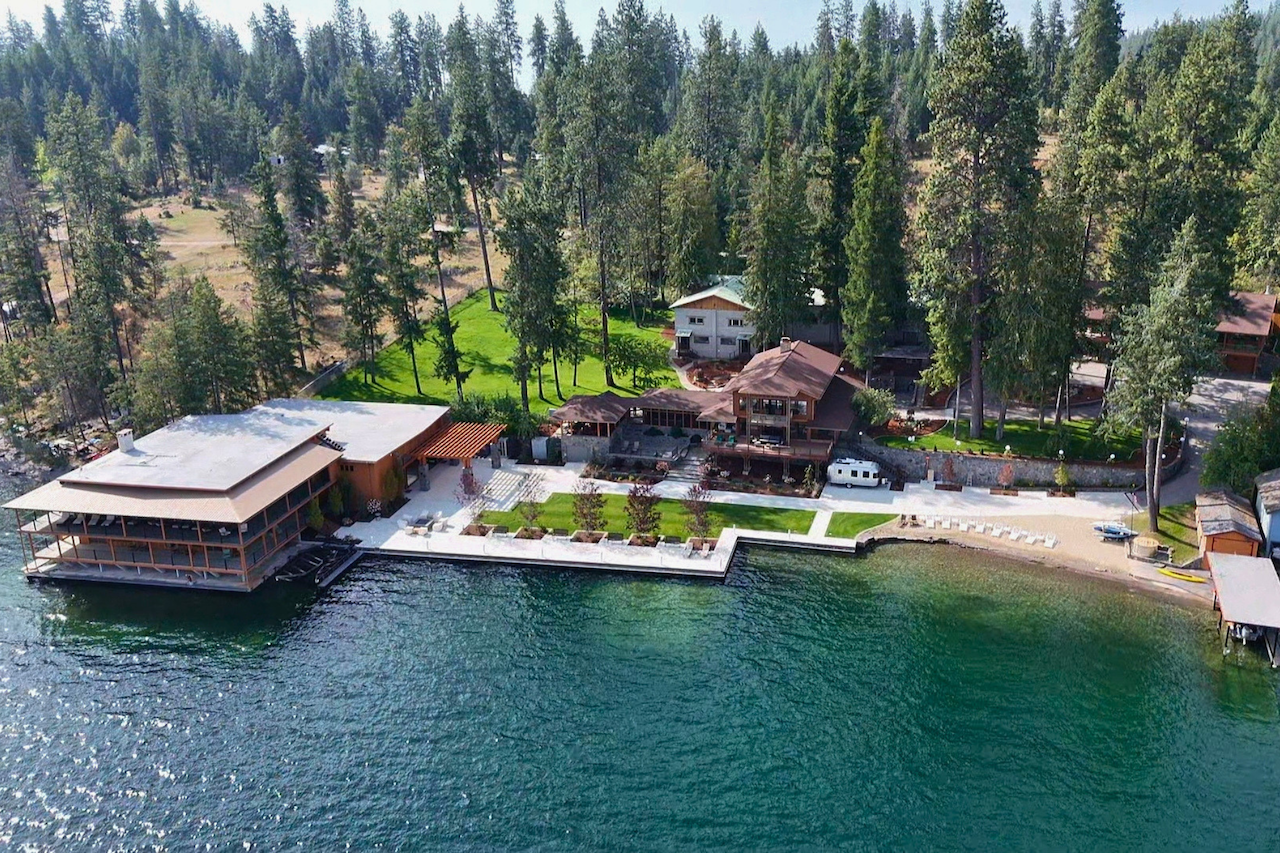 Aerial view of a lakeside property with multiple houses, a large patio, and a boat dock, surrounded by tall trees and forest.