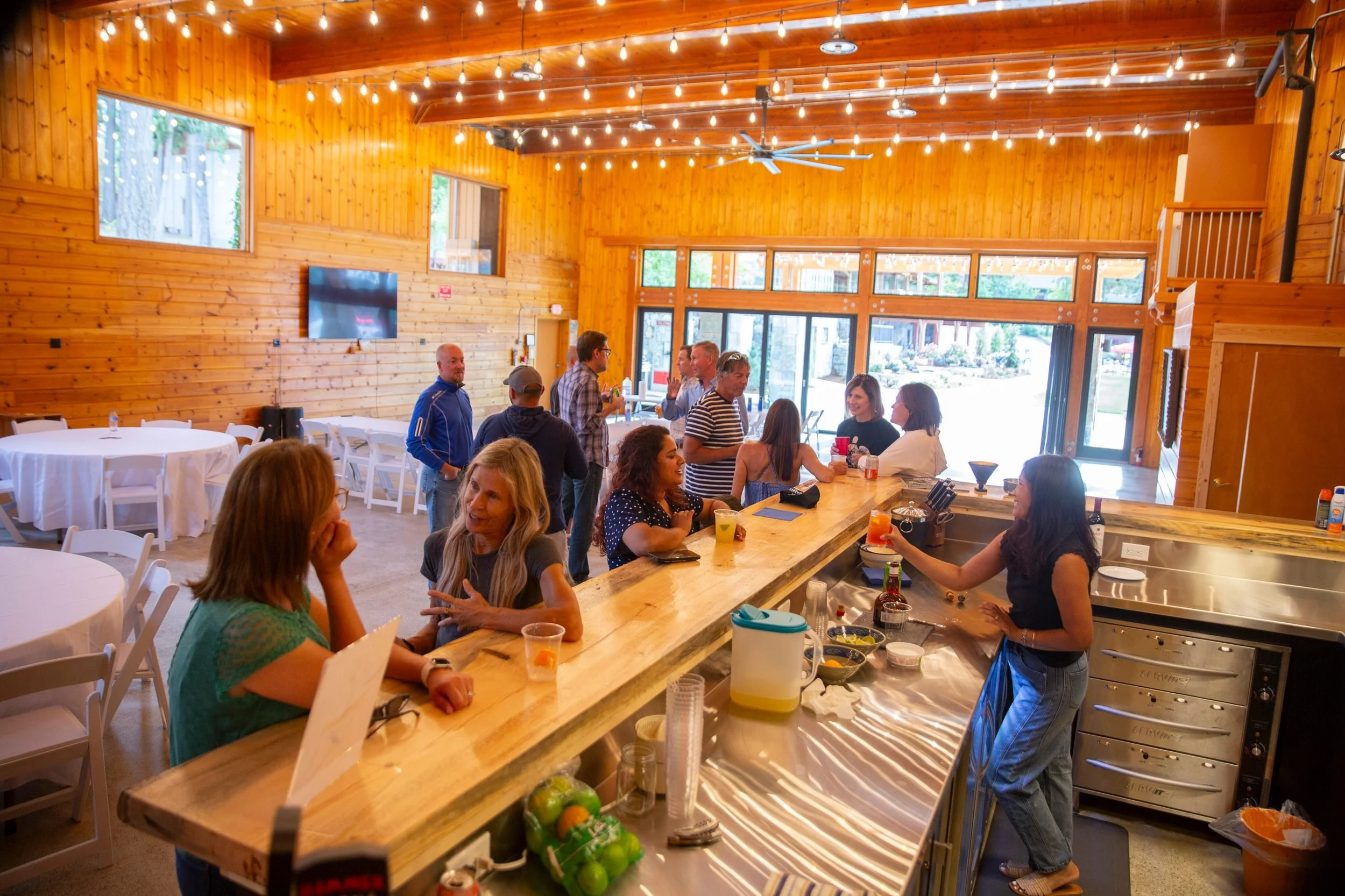 A group of people gathered inside a spacious, warmly lit restaurant with wooden walls and ceiling, talking and enjoying drinks. People are sitting at a long wooden bar and round tables, with some standing and socializing. The restaurant has large windows and string lights hanging from the ceiling.