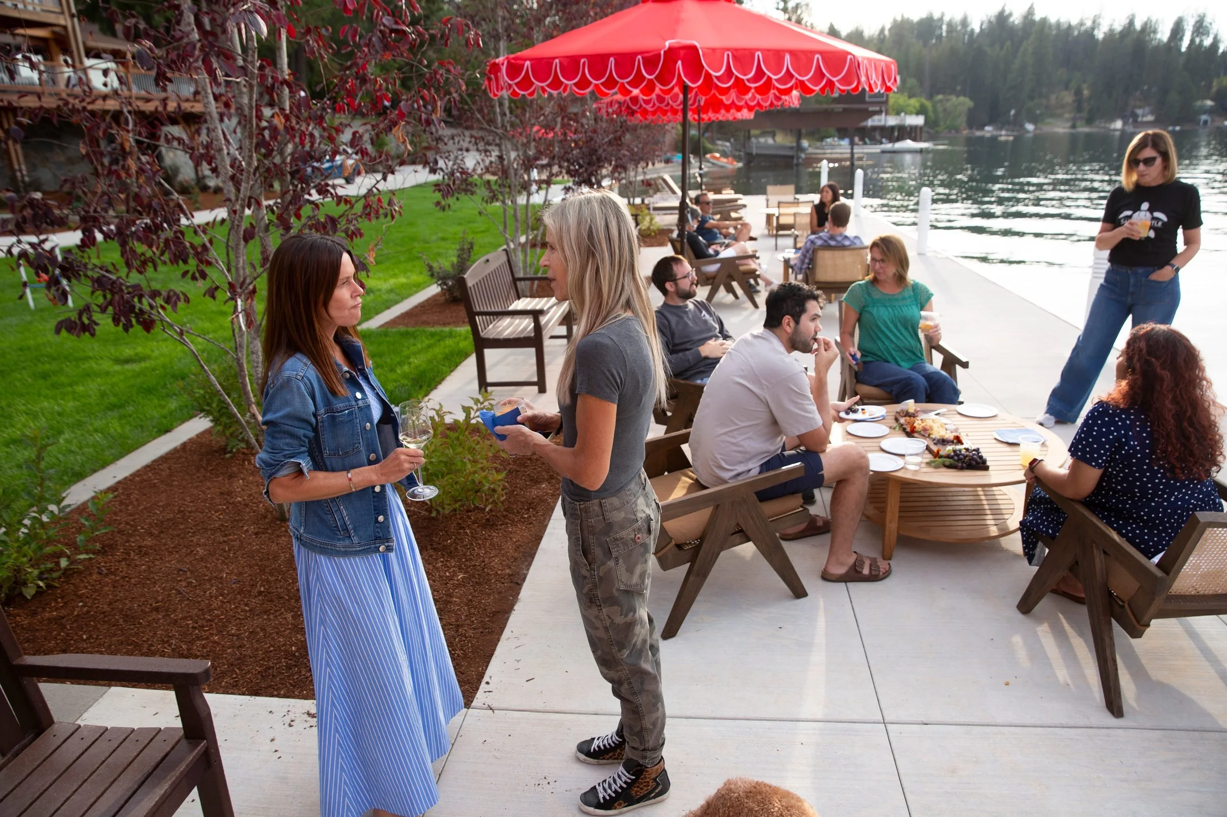 People socializing on a lakeside patio, sitting at tables and standing, with a view of the lake and boats. A red umbrella is providing shade, and the scene appears to be a casual outdoor gathering or party.