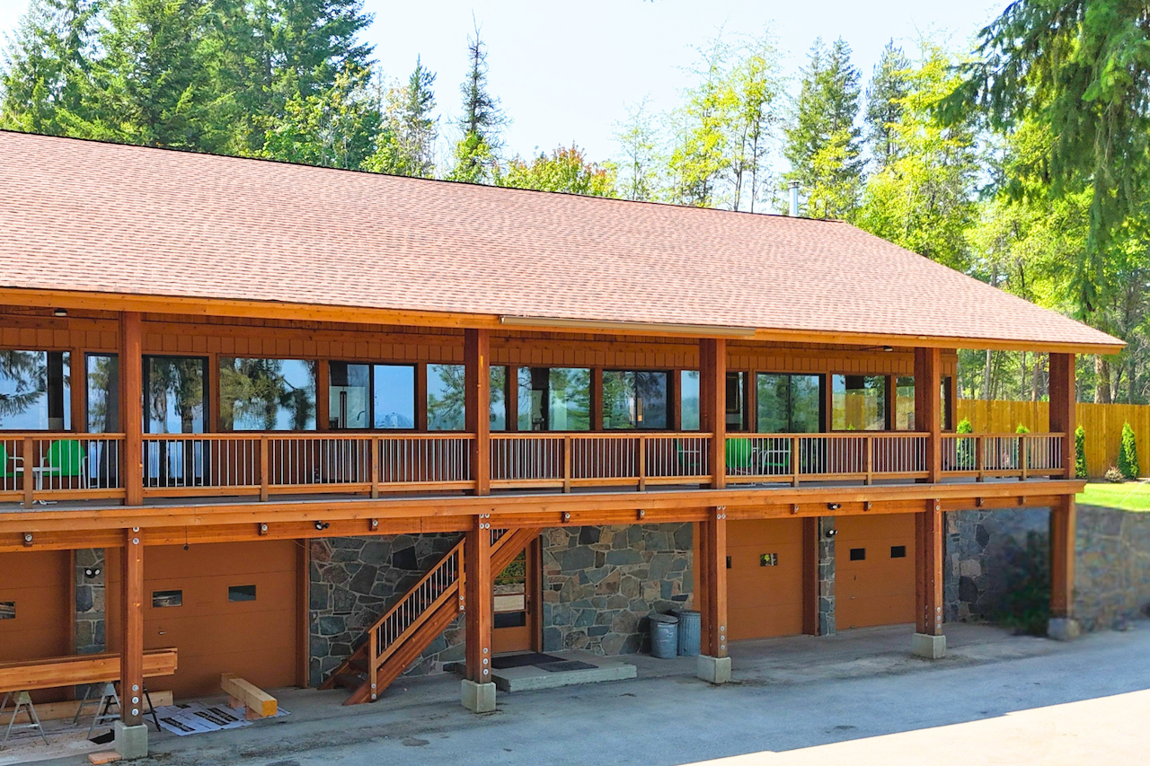 A large wooden house with a porch and four garage doors at the bottom, situated in a wooded area with tall trees and green grass.