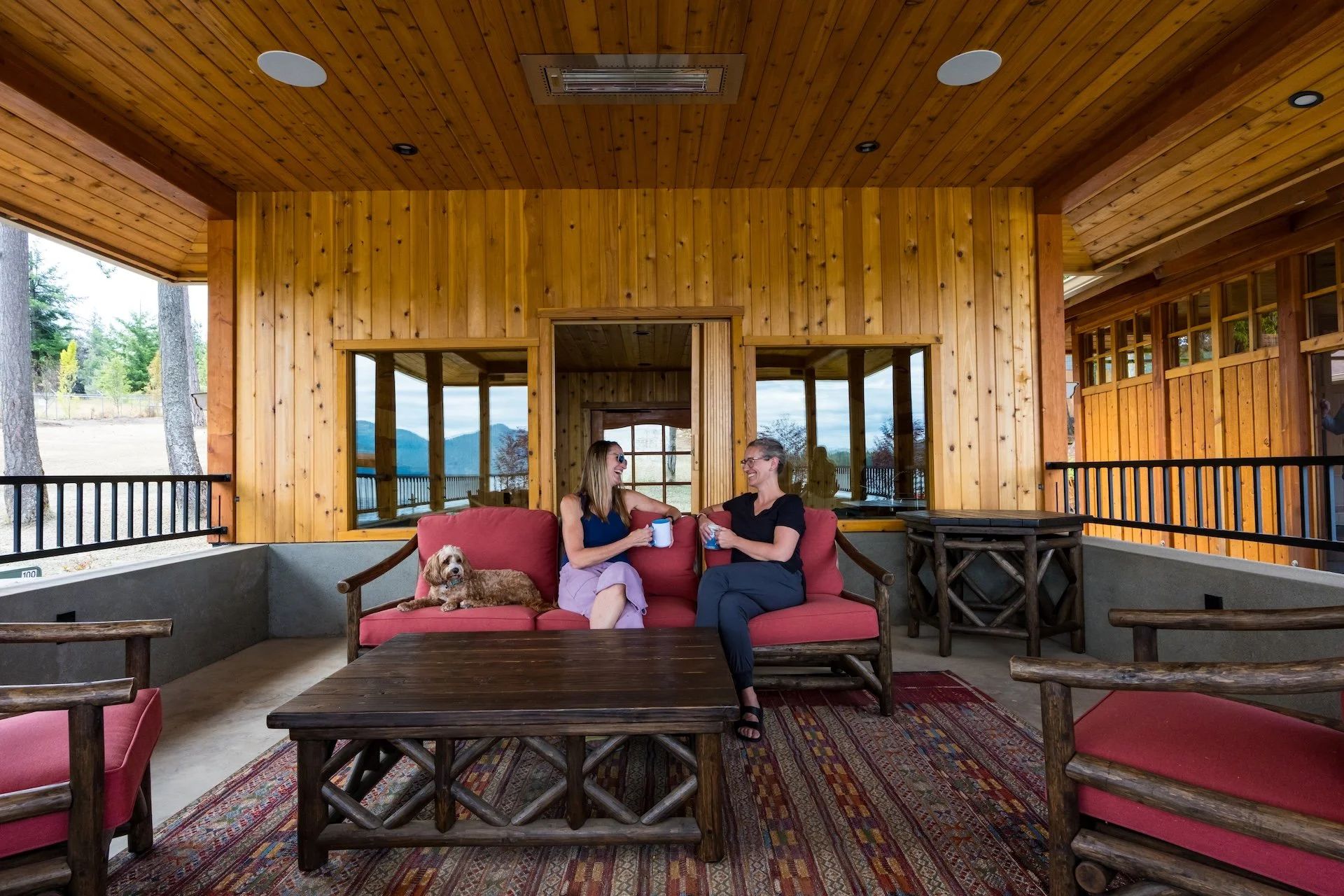 Two women sitting on a red sofa with a dog, holding mugs, in a wooden balcony with mountain and forest views.