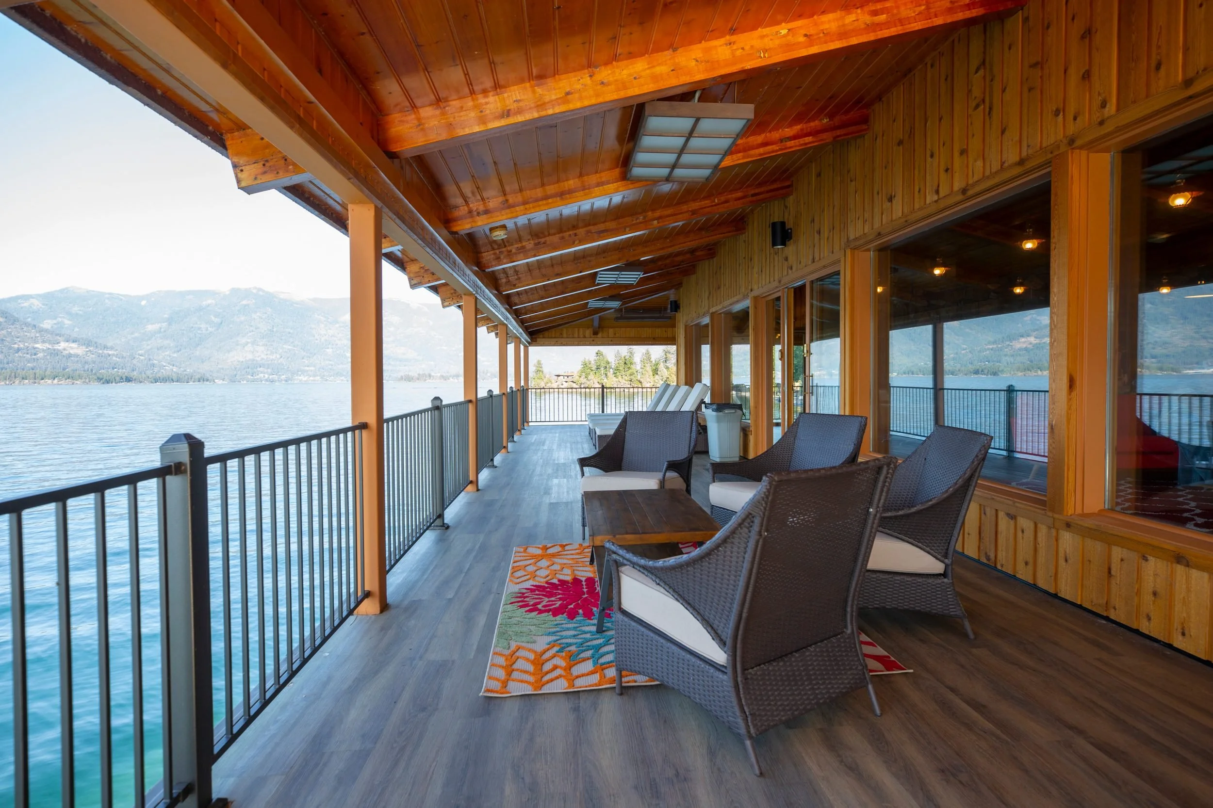 A covered balcony with wooden ceiling and railing overlooks a body of water and distant mountains. The balcony has chairs, a small table, and a colorful rug.