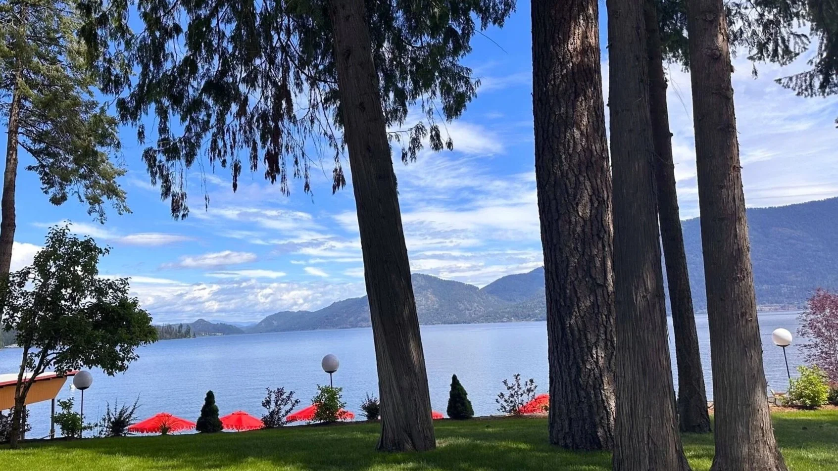 View of a lakeside scene with tall trees in the foreground, a calm blue lake, and mountains in the background under a partly cloudy sky. There are red umbrellas and small trees with spherical lamps on the shore.