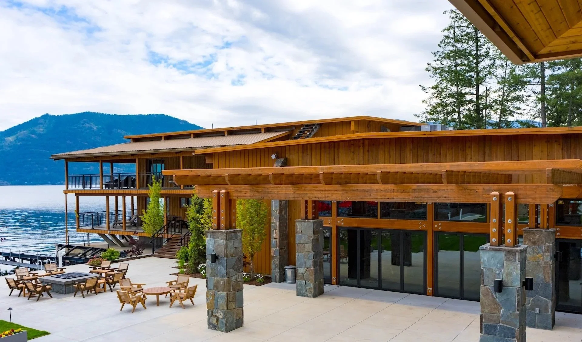 A lakeside building with large windows, wooden and stone exterior, and a patio area with chairs and tables, overlooking a body of water with a mountain in the background, under a cloudy sky.
