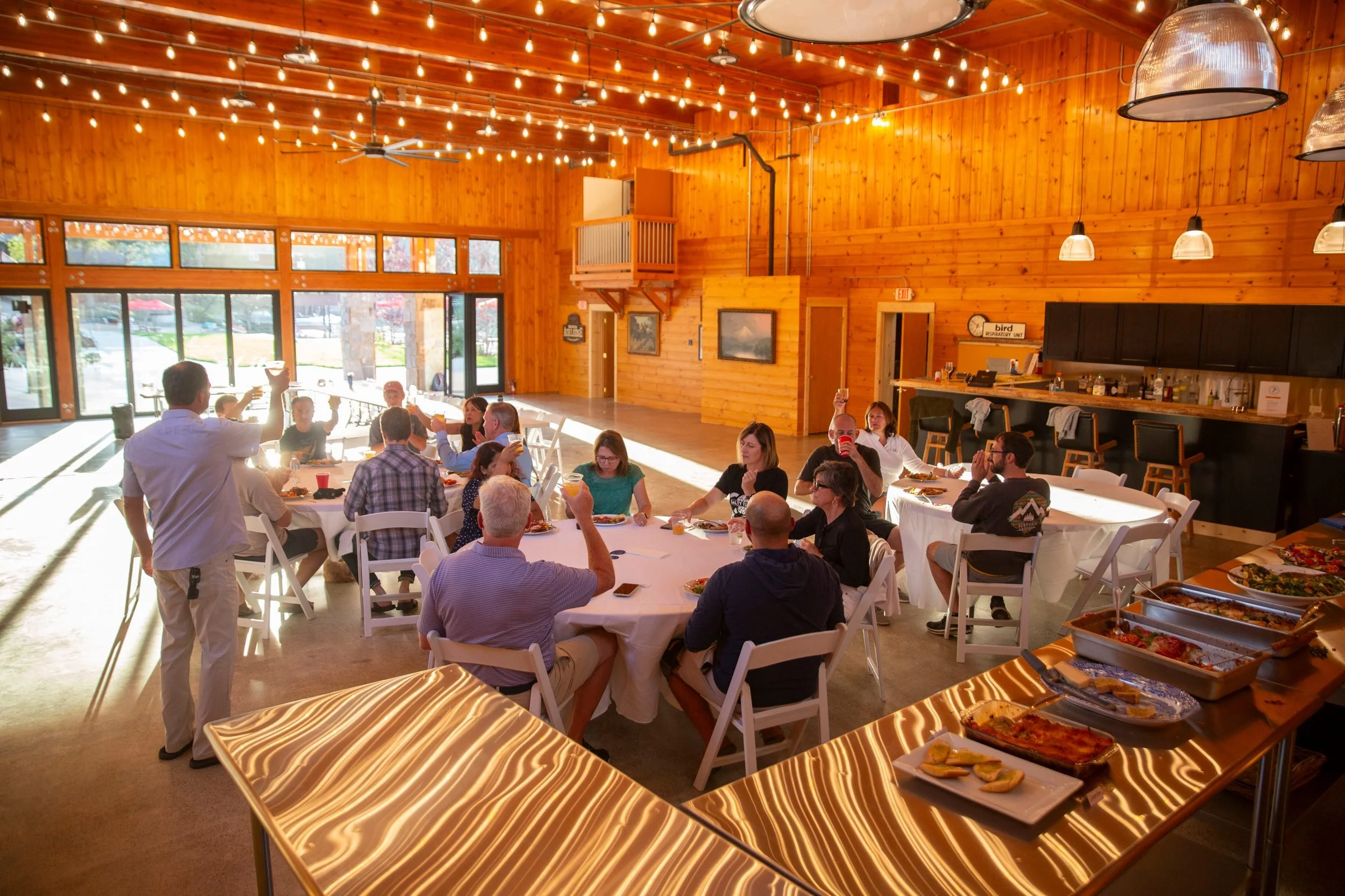 Inside a wood-paneled event hall with large windows, a group of people sit at round tables enjoying a meal and drinks. There are string lights, pendant lights, and a high ceiling with fans. Food trays with pizza and other dishes are on the foreground table.