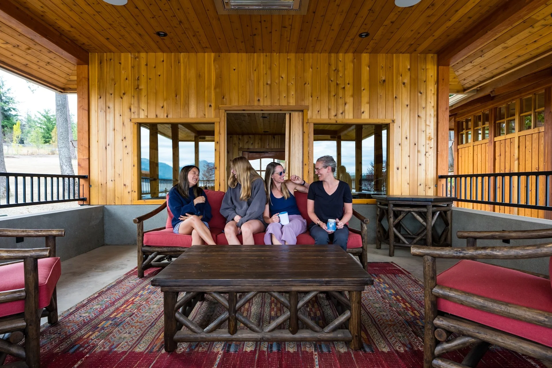 Four women sitting on a red cushioned outdoor sofa, laughing and talking, in front of a wooden building with large windows, inside a covered patio with a colorful rug and wooden furniture.