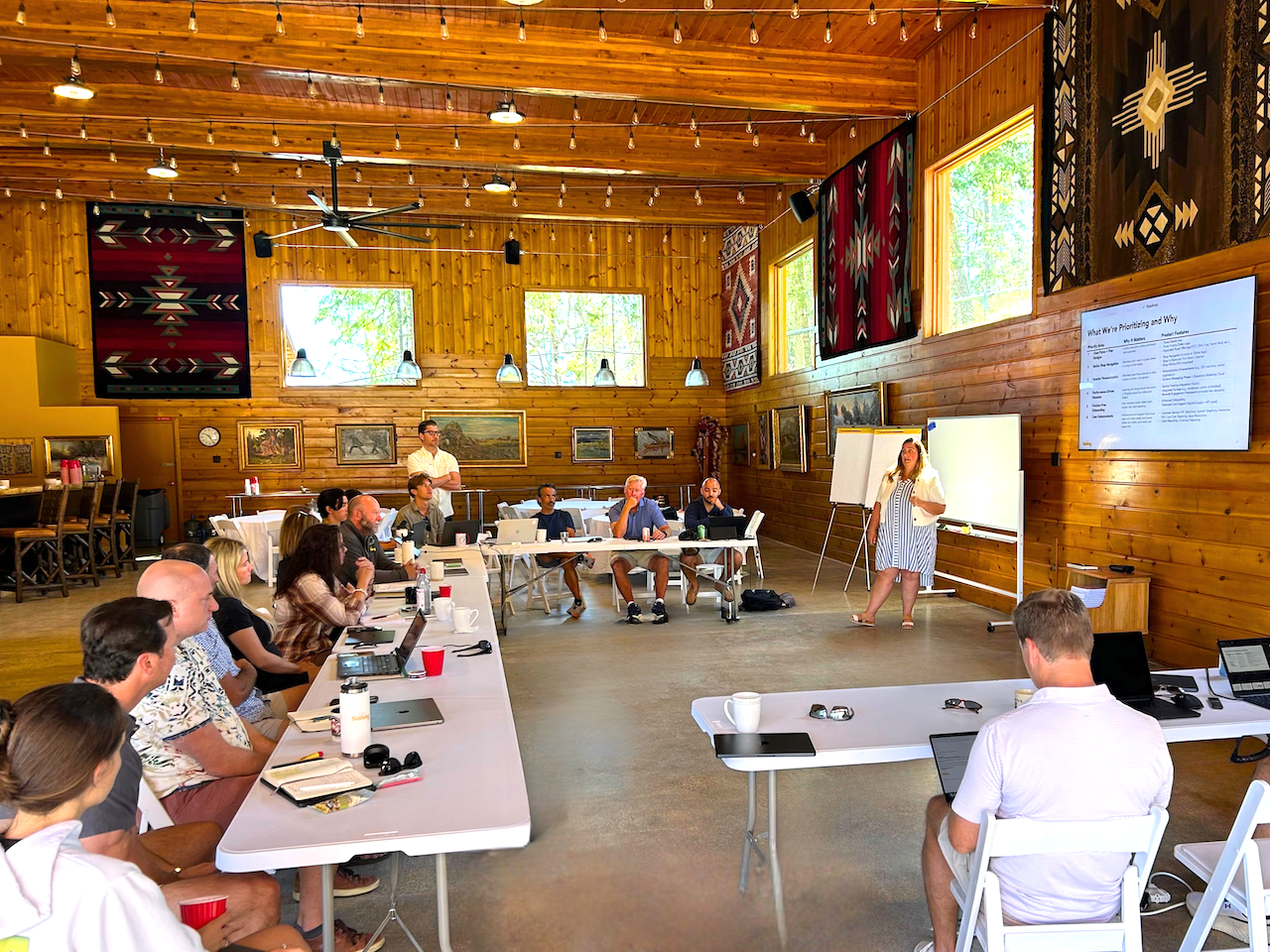 Group of people attending a presentation in a rustic, wood-paneled room with large windows, paintings on the wall, and a woman giving a presentation at a whiteboard