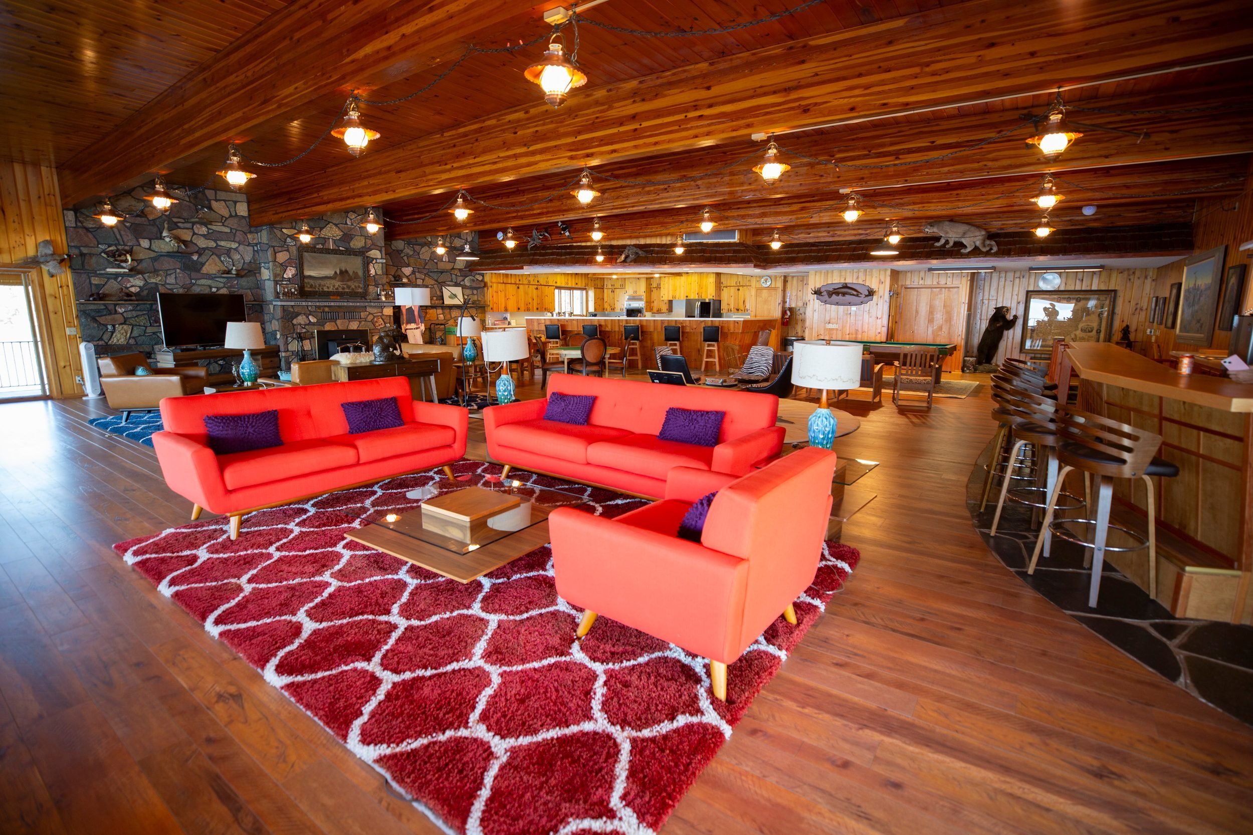 A cozy living room with wooden walls and ceiling, featuring a large stone fireplace, a flat-screen TV, four salmon-colored sofas with dark pillows, and a red patterned rug. There are multiple lamps, some with blue bases, and decorative animal and fish wall-mounted figures, along with a bar area and various chairs and tables.