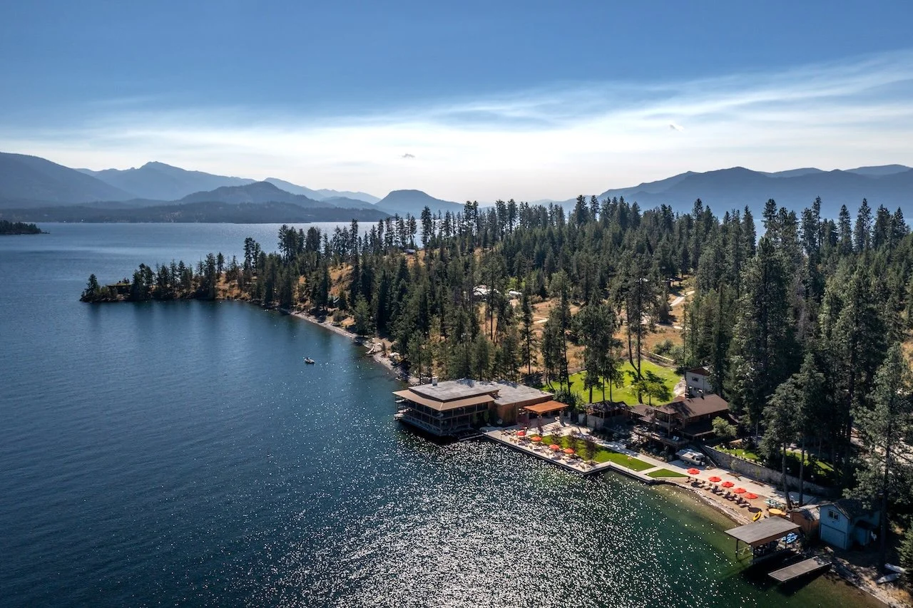 Aerial view of a lakeside resort with buildings, a small beach area with umbrellas, surrounded by pine trees and mountains in the background.