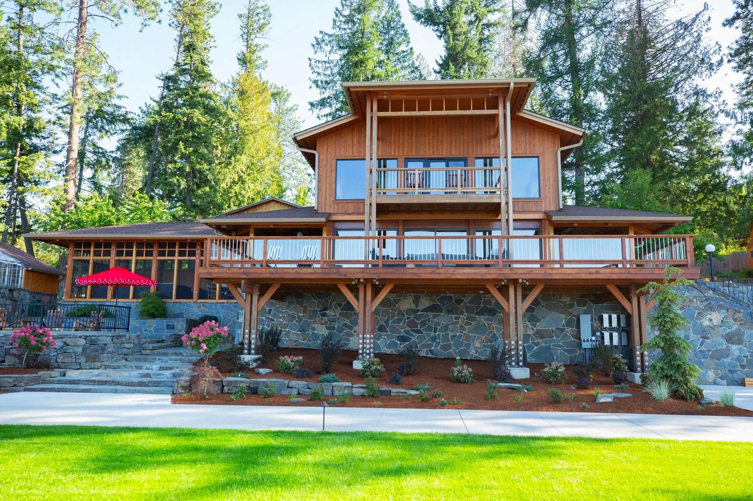 Wooden house with large glass windows, a balcony with a railing, surrounded by trees and a landscaped yard with flowers, shrubs, and a stone retaining wall.