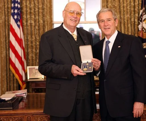 Two men in suits standing in the Oval Office, one of them holding a box with medals or awards inside, with American flags and presidential furnishings in the background.