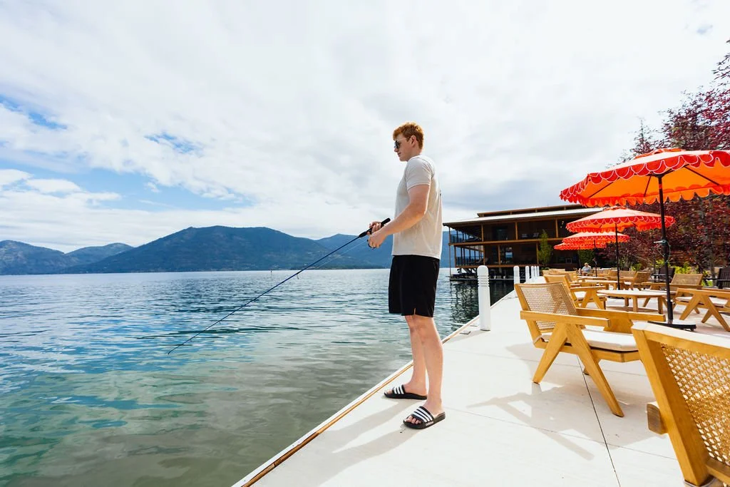 A man in a white shirt, black shorts, and sandals standing on a dock, fishing in a lake with mountain scenery and blue sky in the background, with orange umbrellas and outdoor furniture to the right.