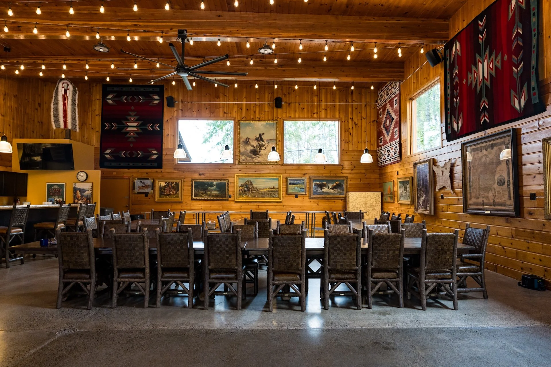 Dining hall with wooden walls and ceiling, decorated with gallery of framed pictures and textiles, large windows with outdoor greenery visible, ceiling fan, string lights, tables and chairs arranged for dining.