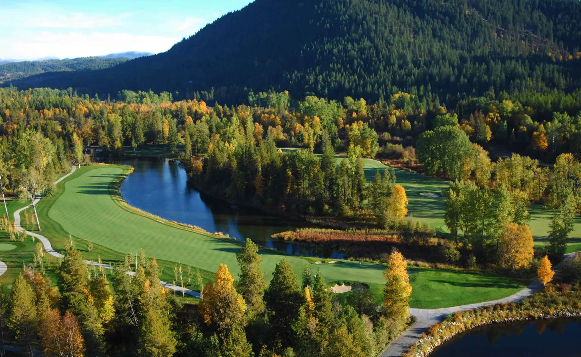 A scenic aerial view of a golf course surrounded by trees with autumn foliage, a river flowing through it, and forested mountains in the background.