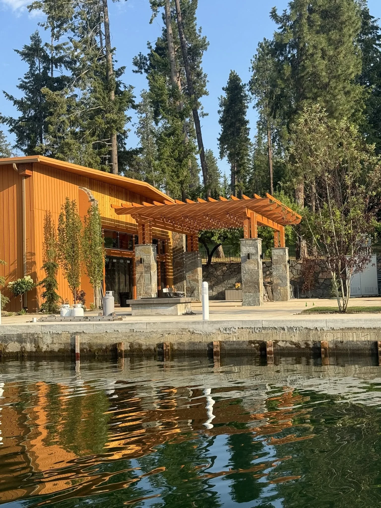 A lakeside house with a wooden pergola, trees, and a calm body of water reflecting the structure and greenery, under a clear blue sky.