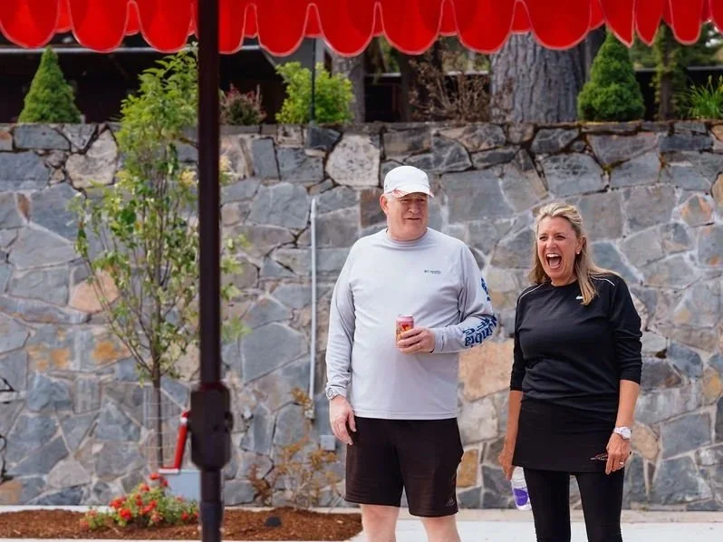 A man and woman standing outdoors at a casual gathering, laughing and talking, with a stone wall and greenery in the background, under a red canopy.