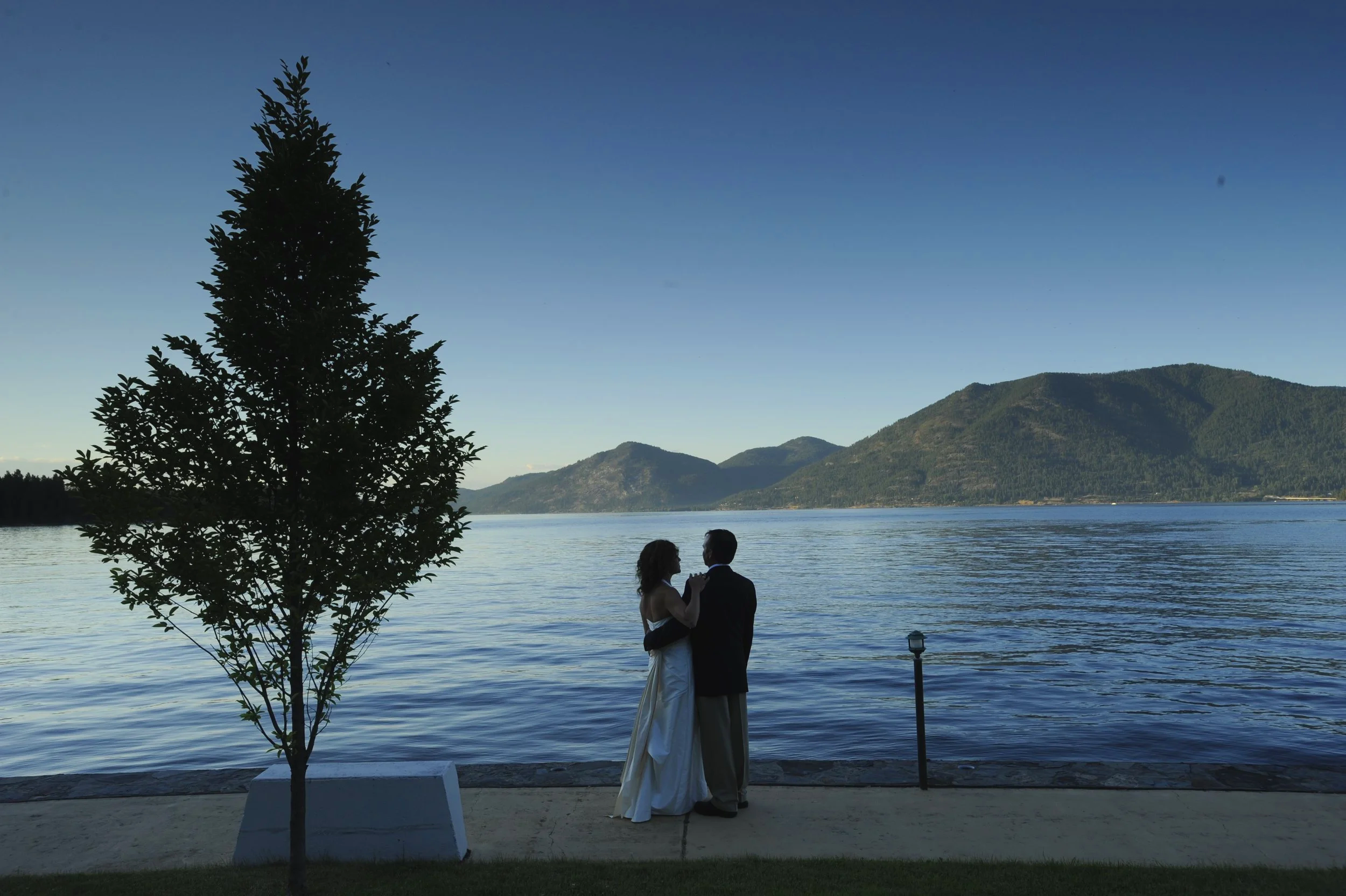 A couple dancing by a lake with mountains in the background, a tree in the foreground, and a lamppost near the water.