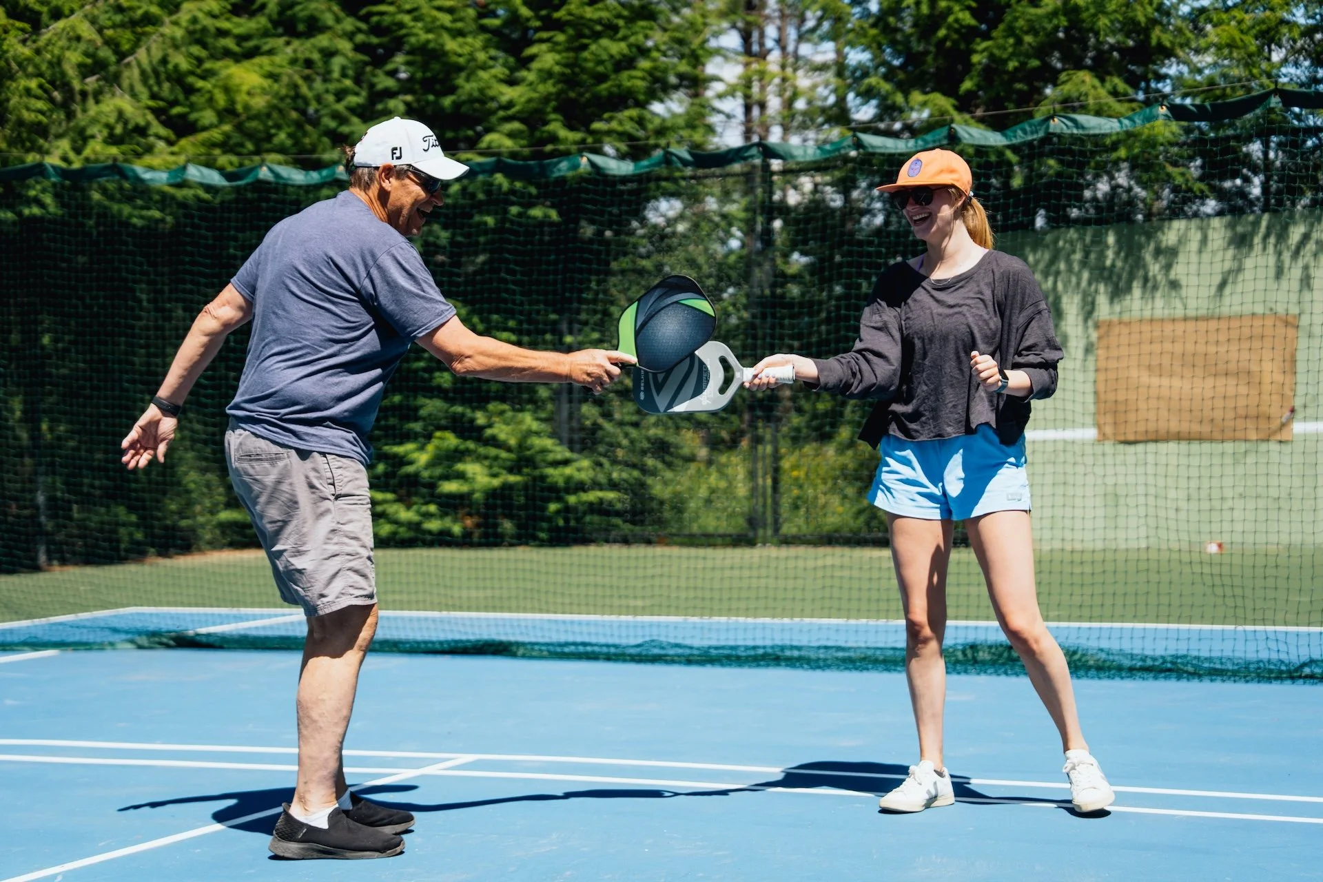 Two people playing pickleball on an outdoor court, with a green net and trees in the background. One person is handing over a paddle to the other, both smiling and wearing casual sportswear and sunglasses.