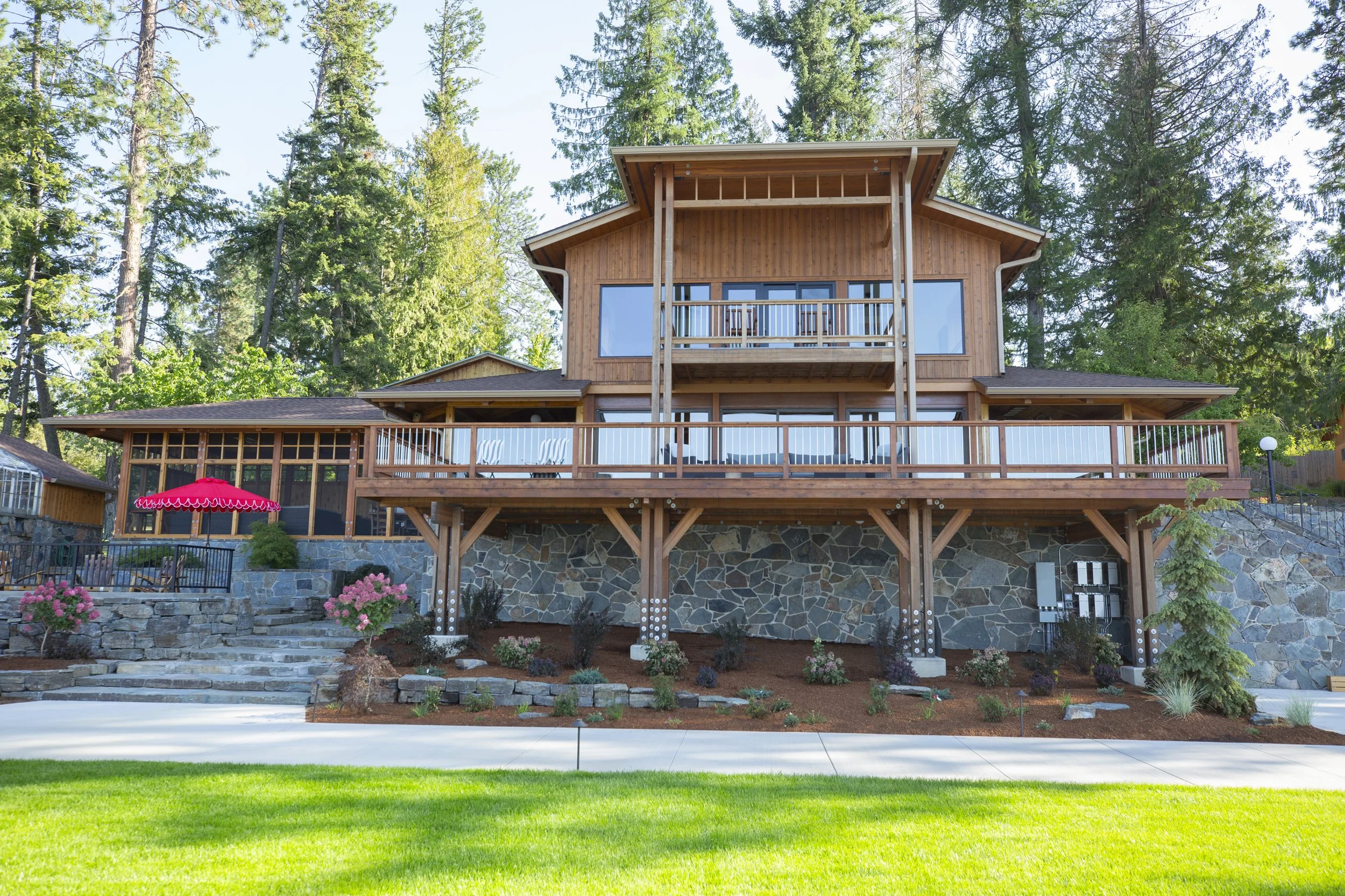 A multi-story wooden house with a large deck, surrounded by trees, with a garden below featuring flowering plants and shrubs.
