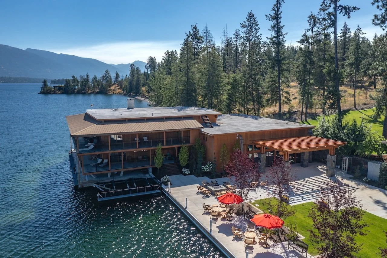 A large house by a lake with outdoor seating, red umbrellas, trees, and mountains in the background.