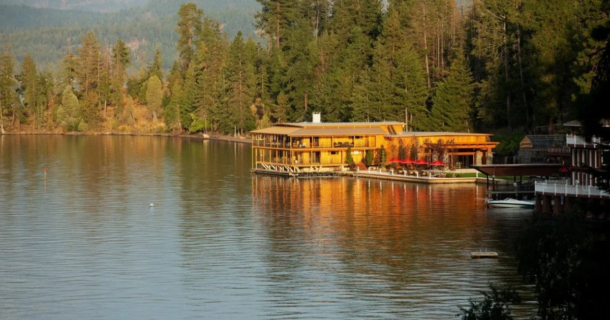 A lakeside scene with a large yellow building and a dock, surrounded by tall pine trees during sunset.