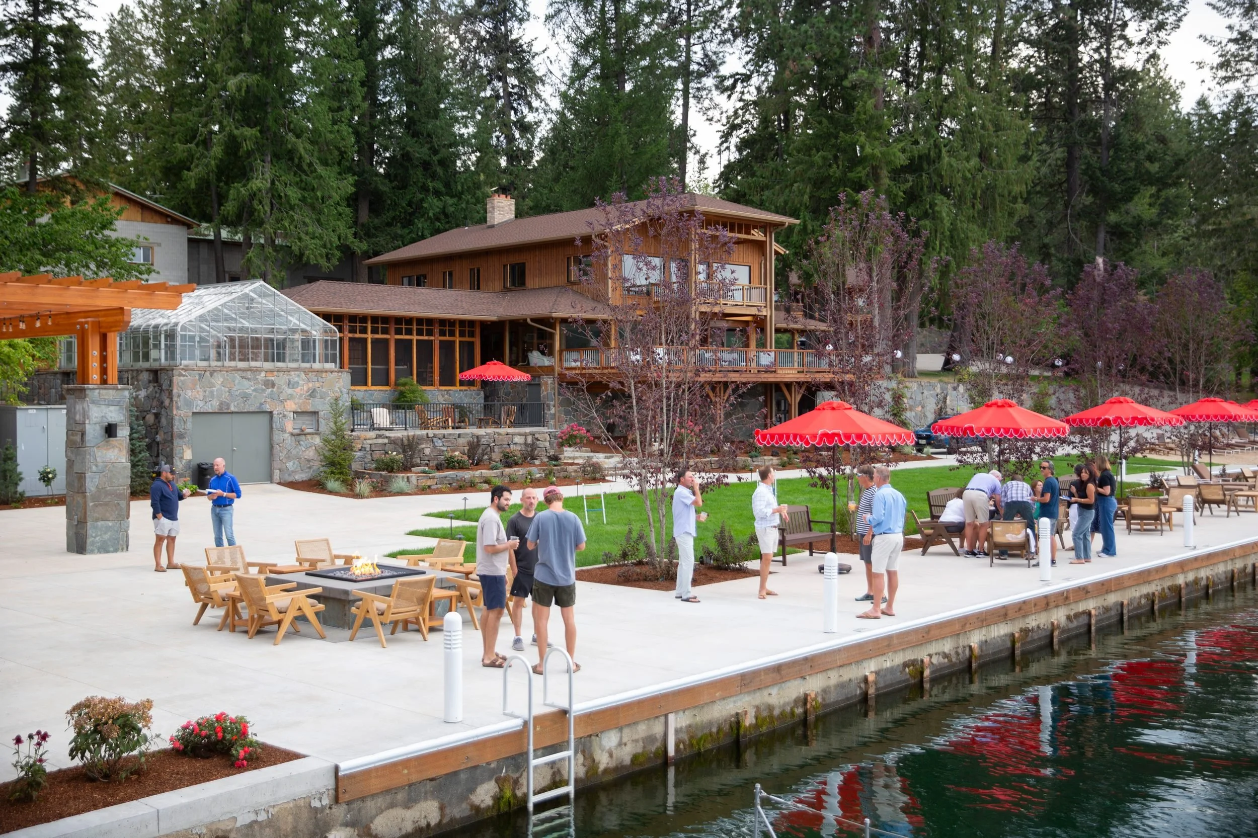 People gathered at a waterfront outdoor event with a large wooden house and lush green trees in the background.
