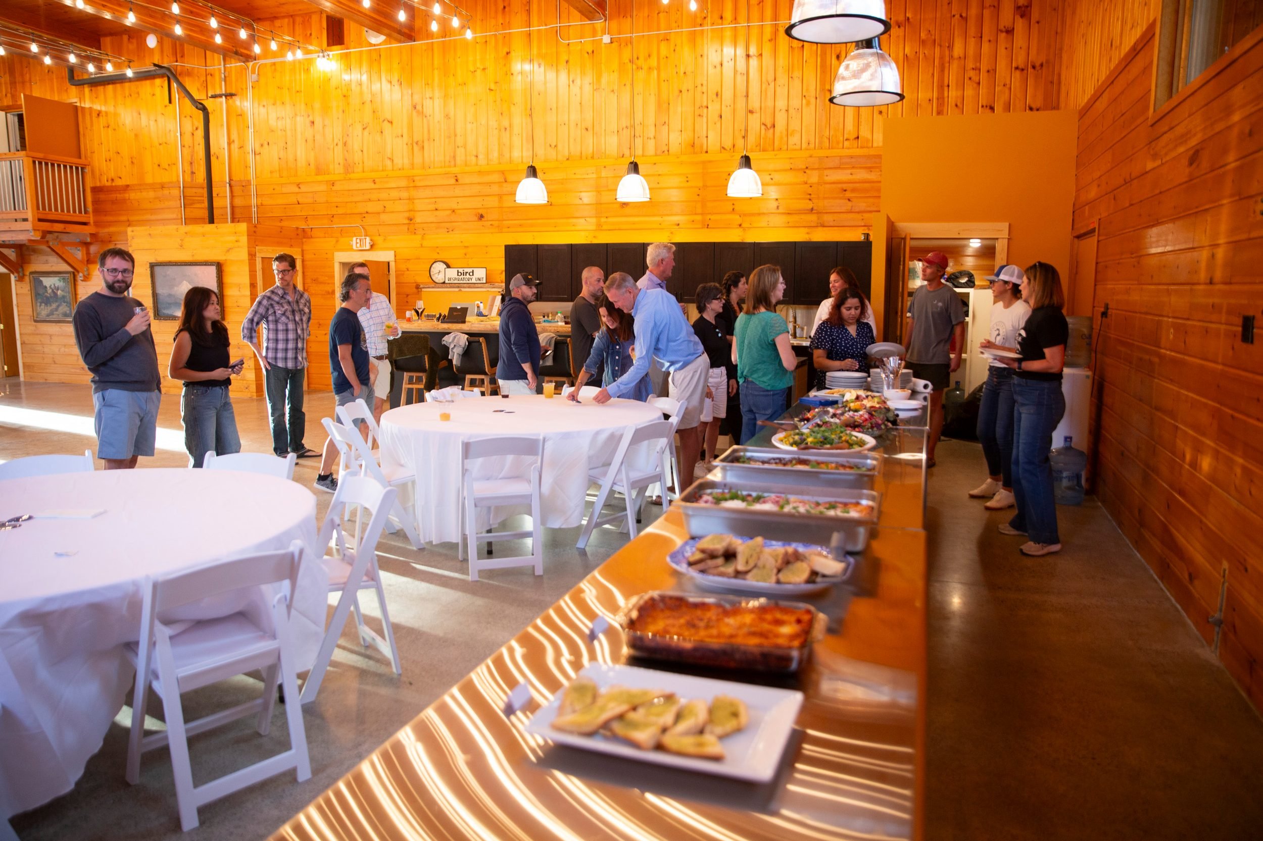 People gathered around a buffet table with food in a wooden-paneled banquet hall with string lights.