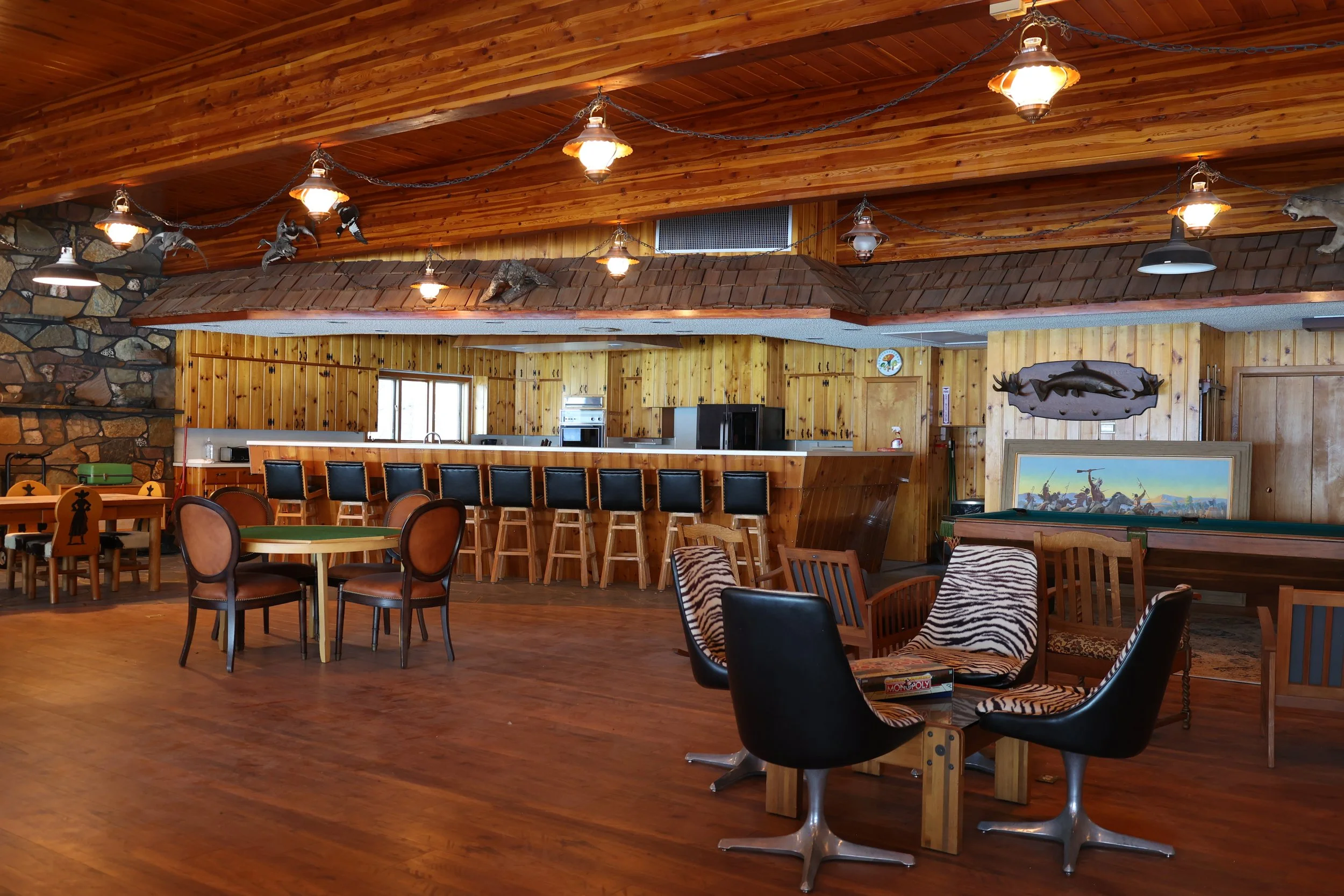 Interior of a cabin-style lodge or restaurant with wood paneled walls and ceiling, rustic lighting fixtures, and a bar area with chairs. Decorated with mounted animals and a painting, featuring a mix of tables and chairs.