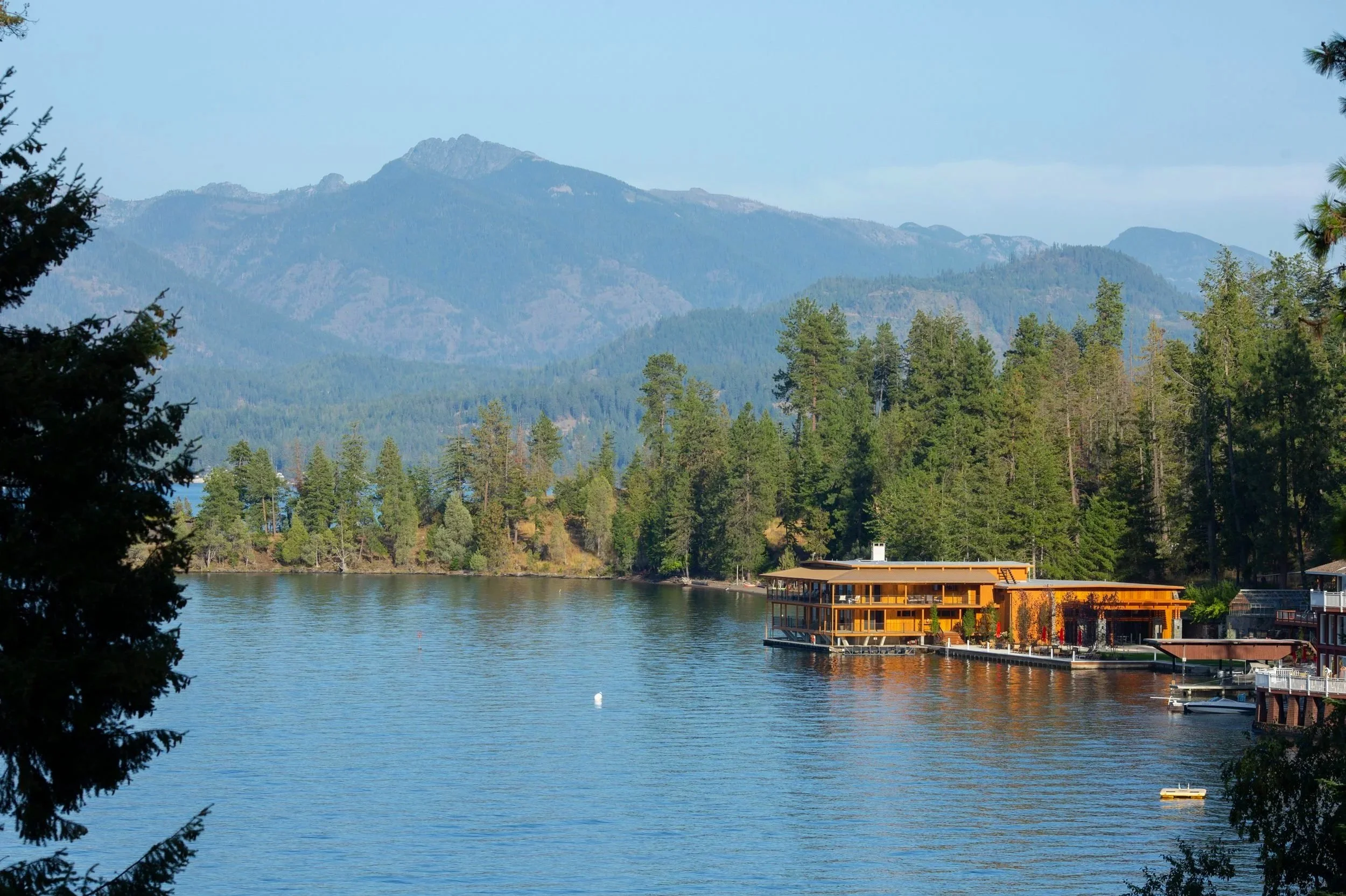A lake surrounded by tall green trees with mountains in the background under a clear blue sky. A modern building with large windows and a deck extends over the water.