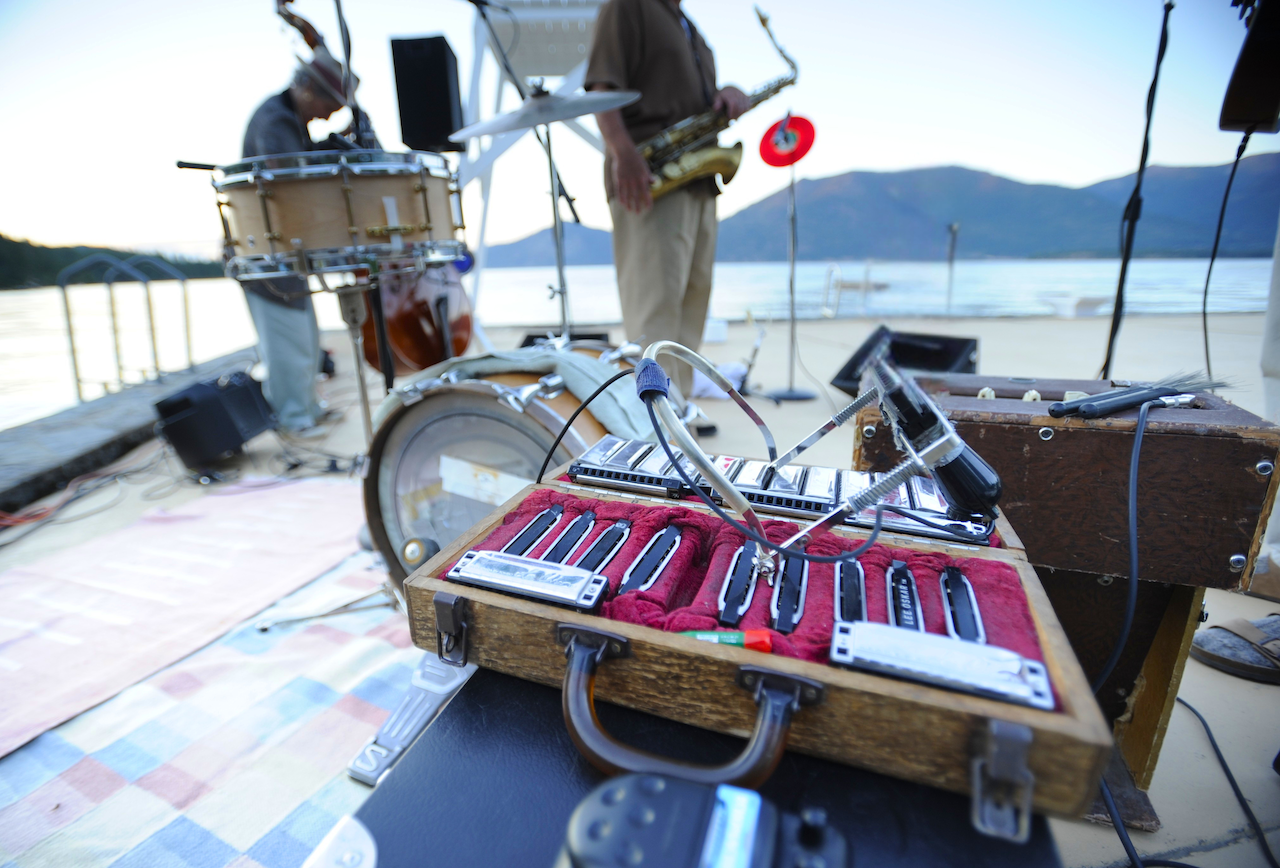 Musicians playing instruments on a lakeside stage with a scenic mountain backdrop.
