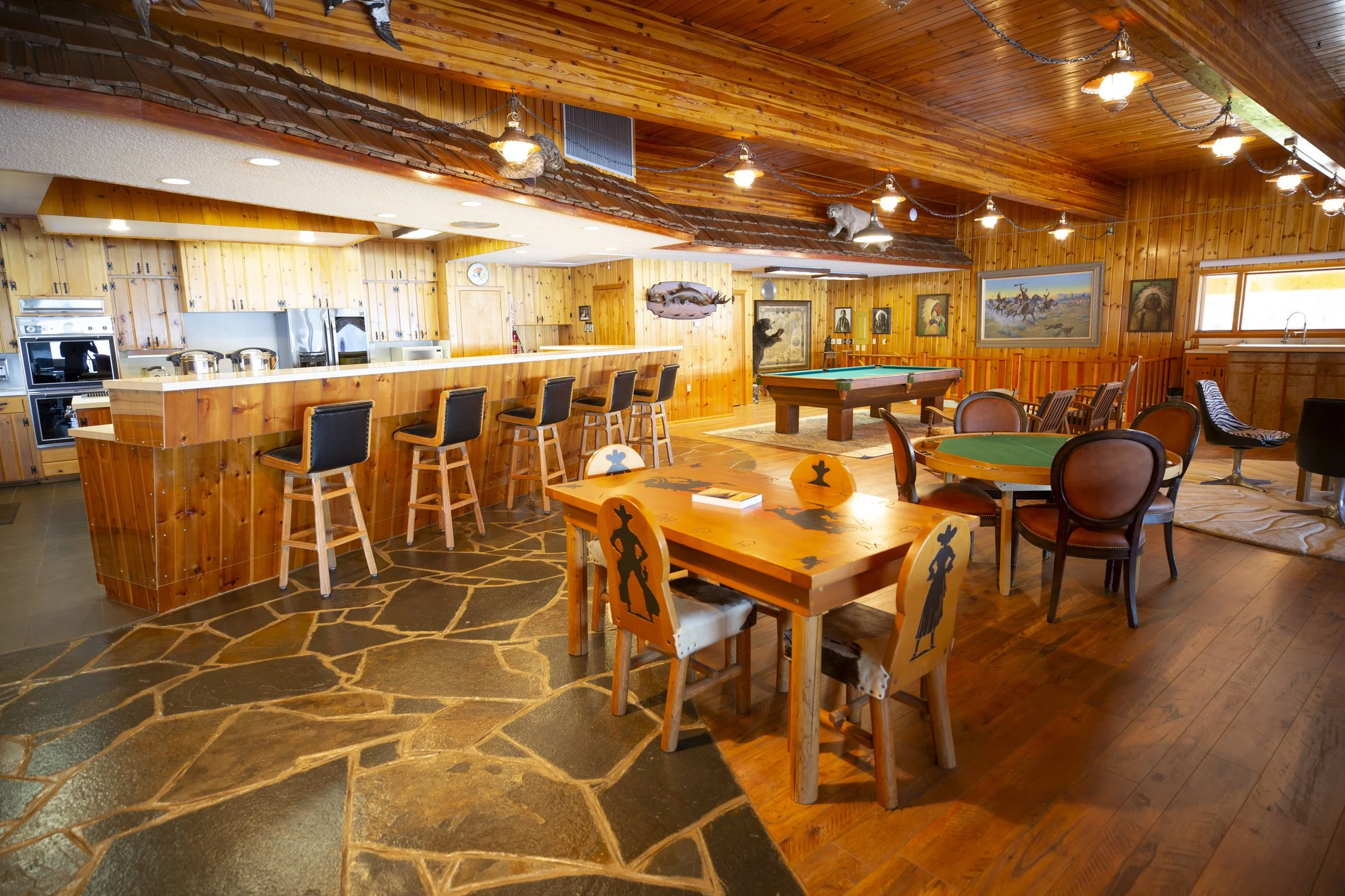 Interior of a rustic lodge game room with wooden walls and ceiling, featuring a pool table, dining tables with chairs, a bar area with stools, and various hunting and fishing themed decorations, including paintings and taxidermy.