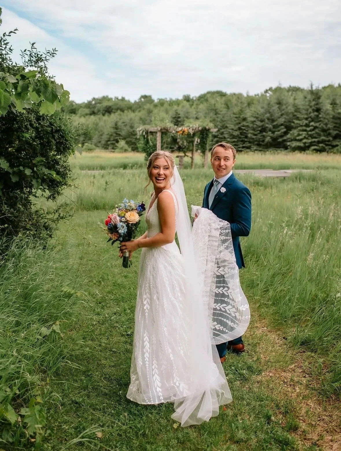 Bride and groom outdoors in a green field with trees, smiling and holding a bouquet of flowers, on their wedding day.
