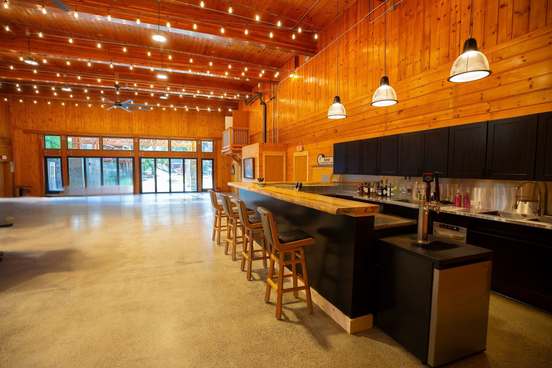Spacious indoor community space with wooden walls and ceiling, warm lighting, a bar counter with barstools, and large windows at the far end showing an outdoor view.